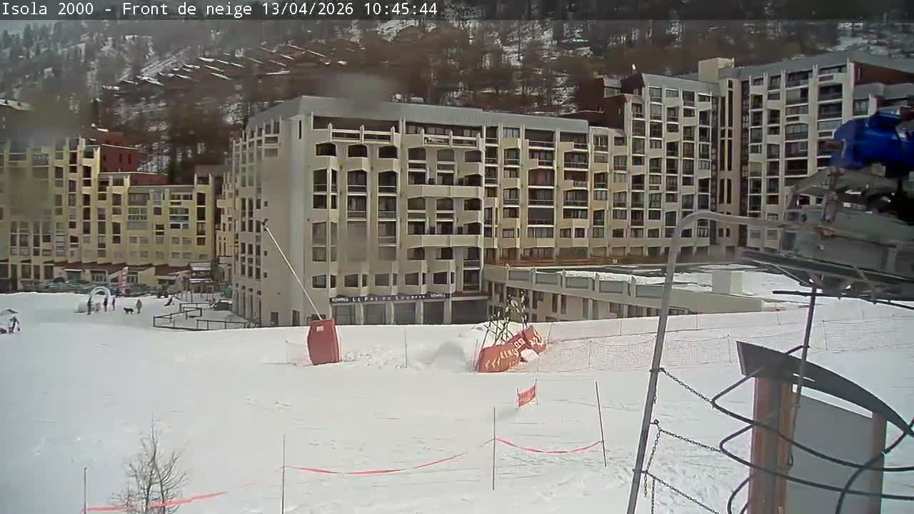 A deserted, snow-covered ski resort area with large buildings and scattered trees is visible, marked by red poles and a barrier tape, all under an overcast winter sky.