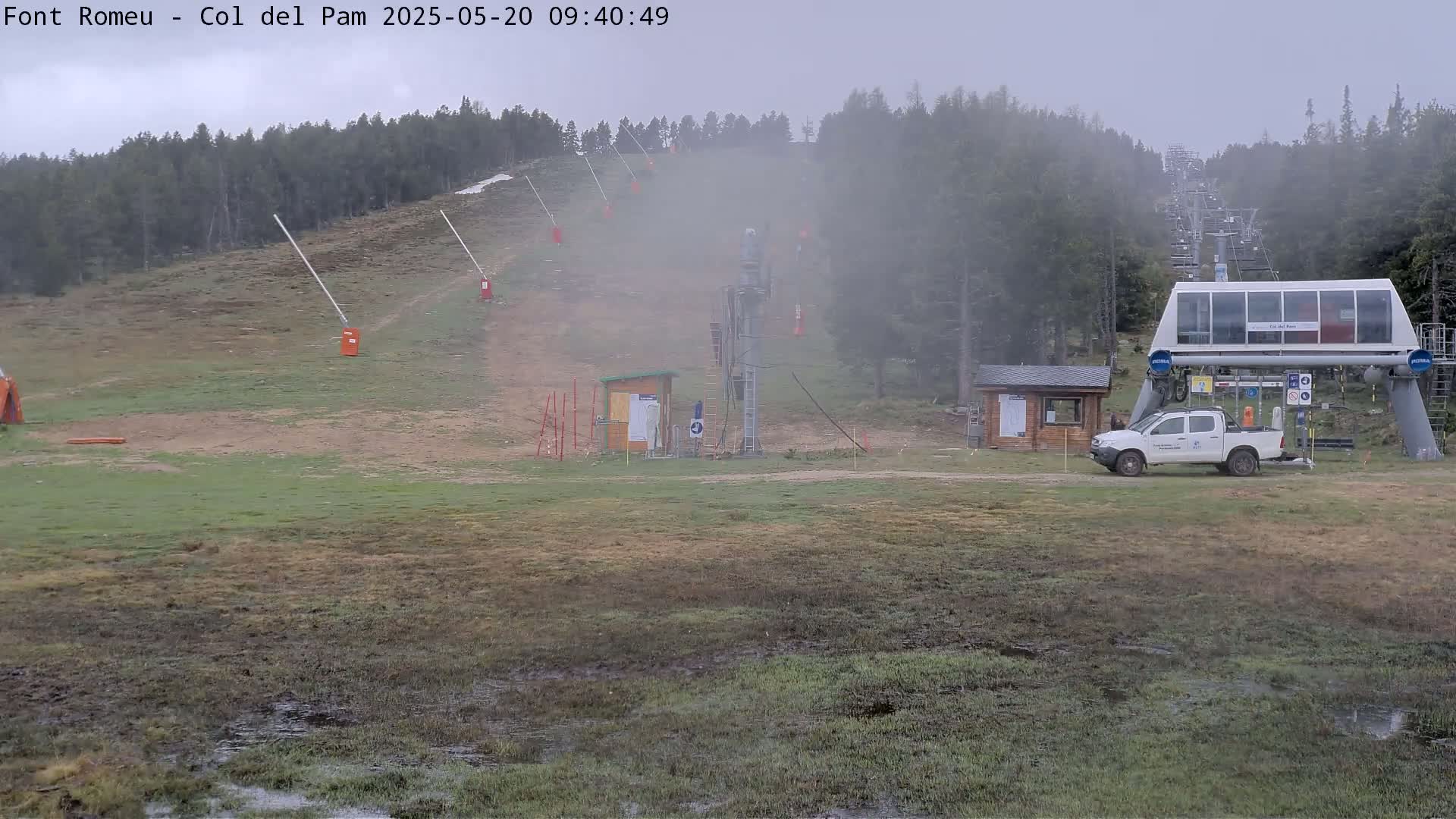A foggy, overcast day at a ski lift base area shows a chairlift ascending a hill lined with trees, a small pickup truck parked near a building, and puddles of water on the wet grassy ground.