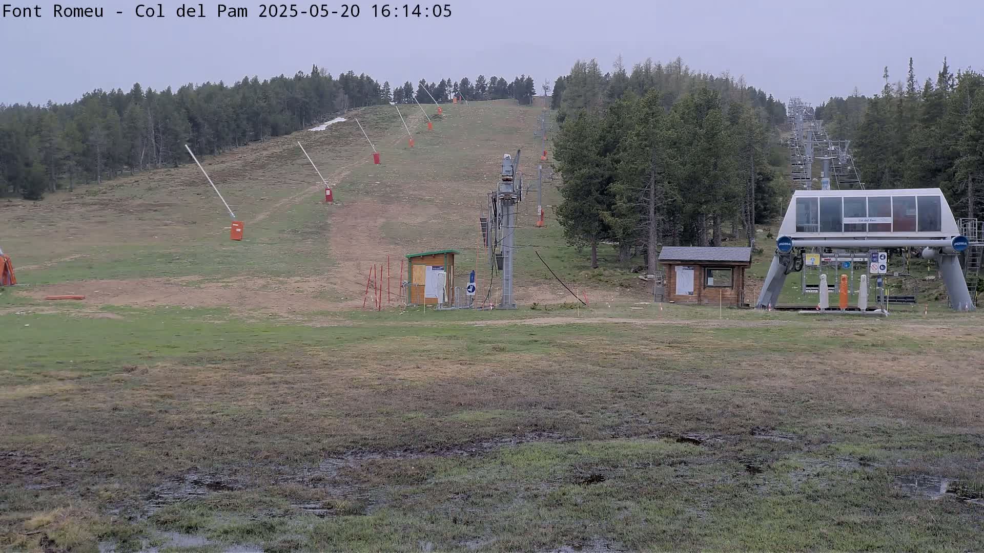 A chairlift base station sits at the bottom of a grassy ski slope under an overcast sky, with muddy patches visible in the foreground.