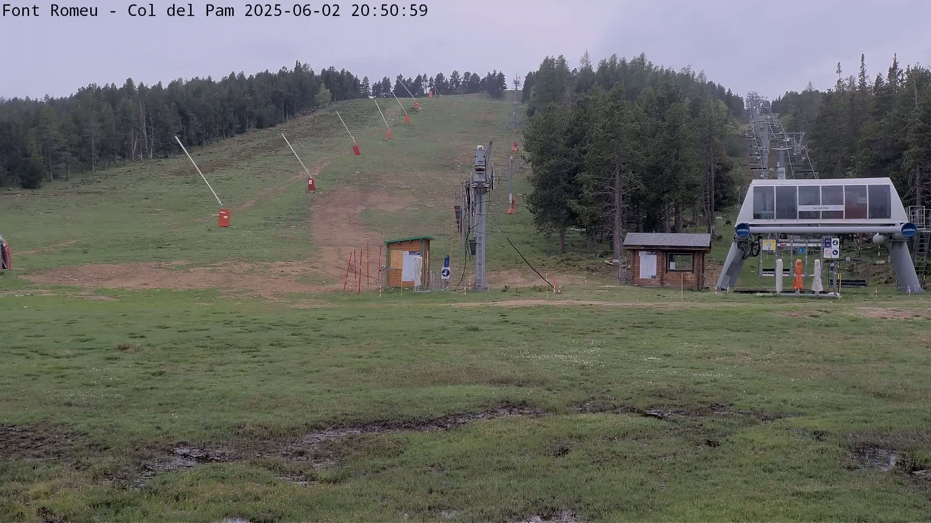 A ski lift base station sits in a grassy field with muddy patches, under an overcast sky, surrounded by a forest.
