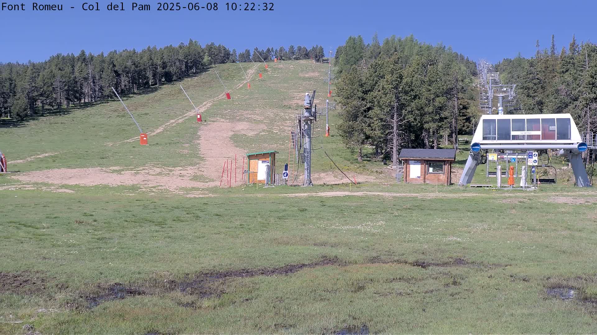 A ski lift base station sits on a grassy hillside under a clear blue sky, with a ski lift ascending a hill covered in sparse vegetation.