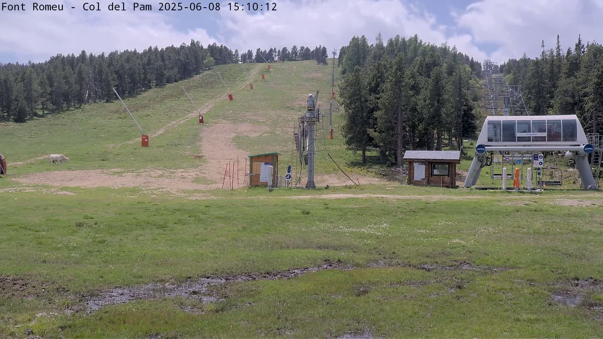 A chairlift base station sits in a grassy field with a cow grazing nearby, under a partly cloudy sky.