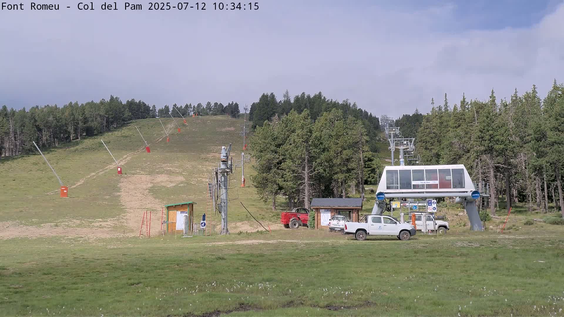 A ski lift base station with several vehicles is situated on a grassy hill, surrounded by evergreen trees under a mostly clear sky.