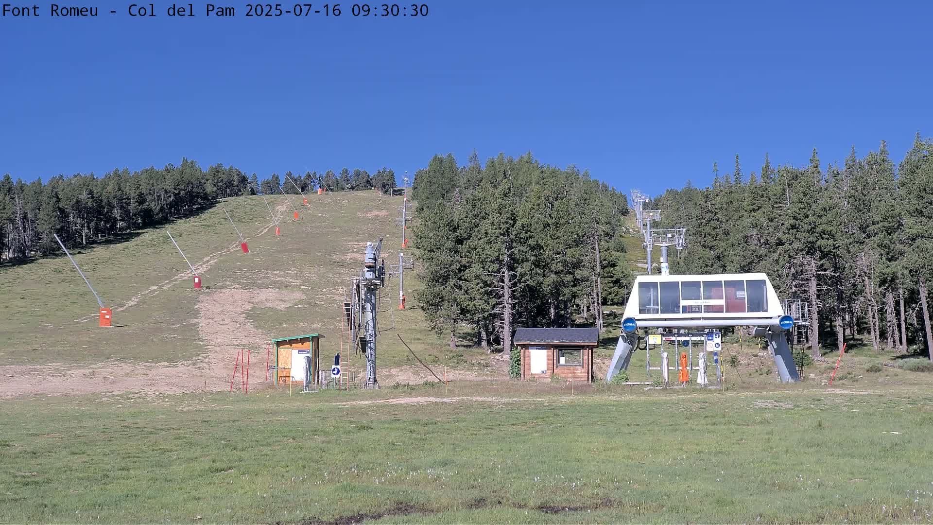 A ski lift base station sits in a grassy area surrounded by a coniferous forest under a clear blue sky.