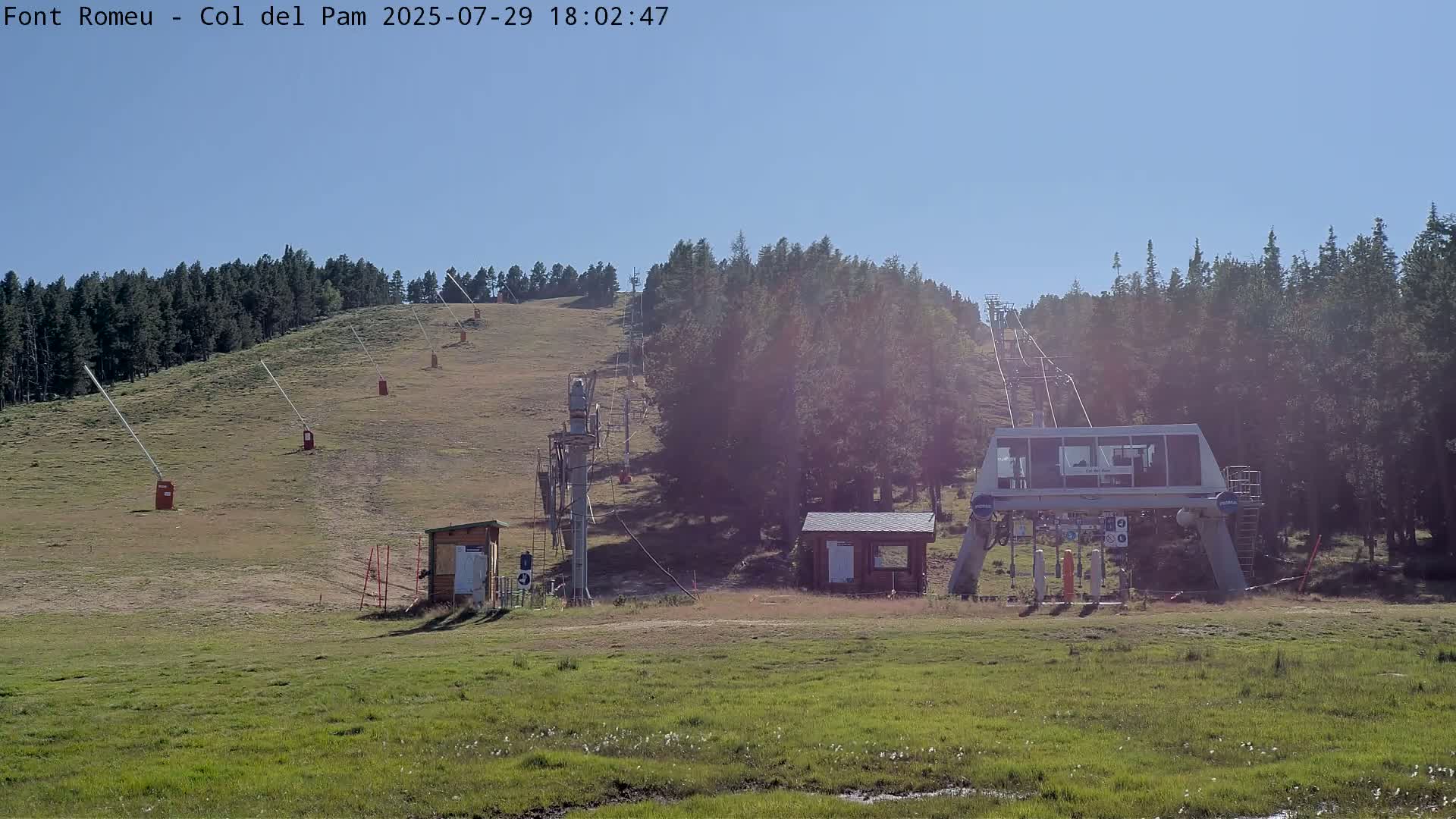 A ski lift base station sits in a grassy field at the bottom of a hill covered in pine trees under a clear blue sky.