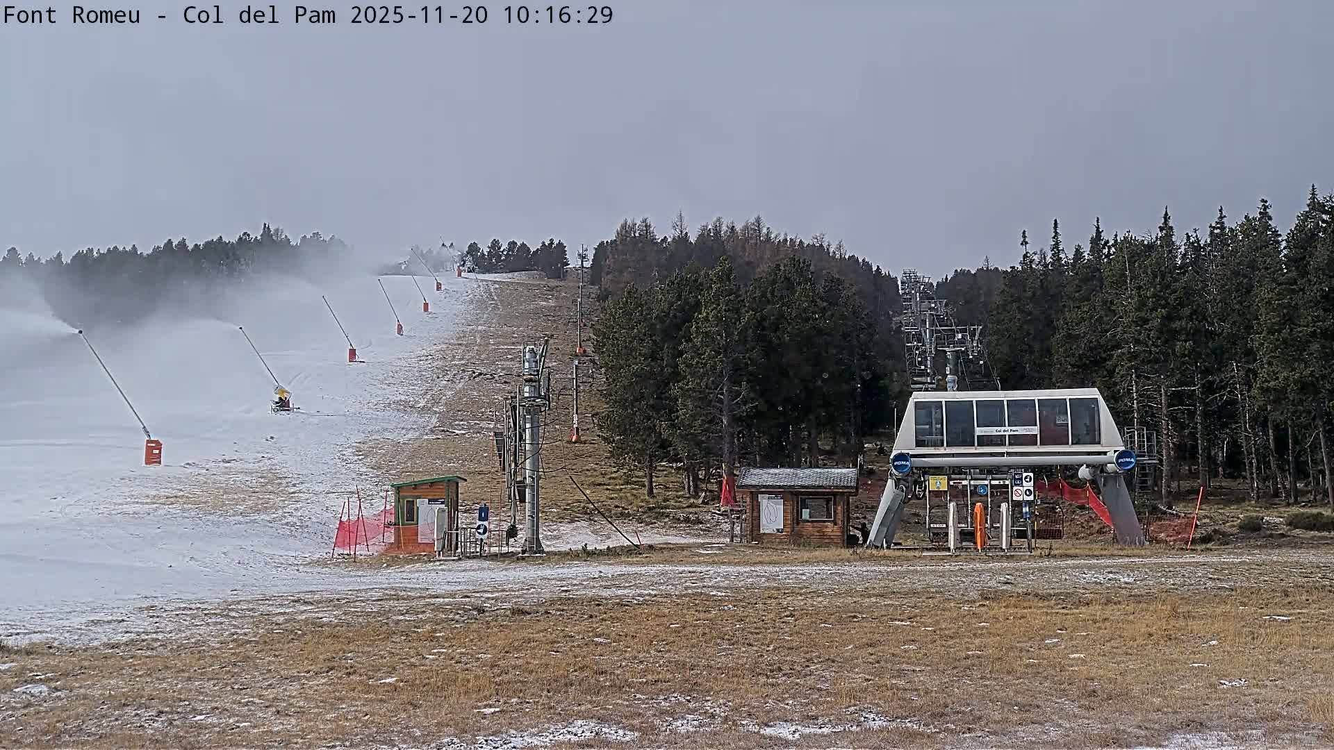 On an overcast day, a ski slope is actively being prepared with snow cannons spraying snow across a partially covered, dry grassy hill, leading up to a prominent chairlift station and ascending line amidst evergreen trees.