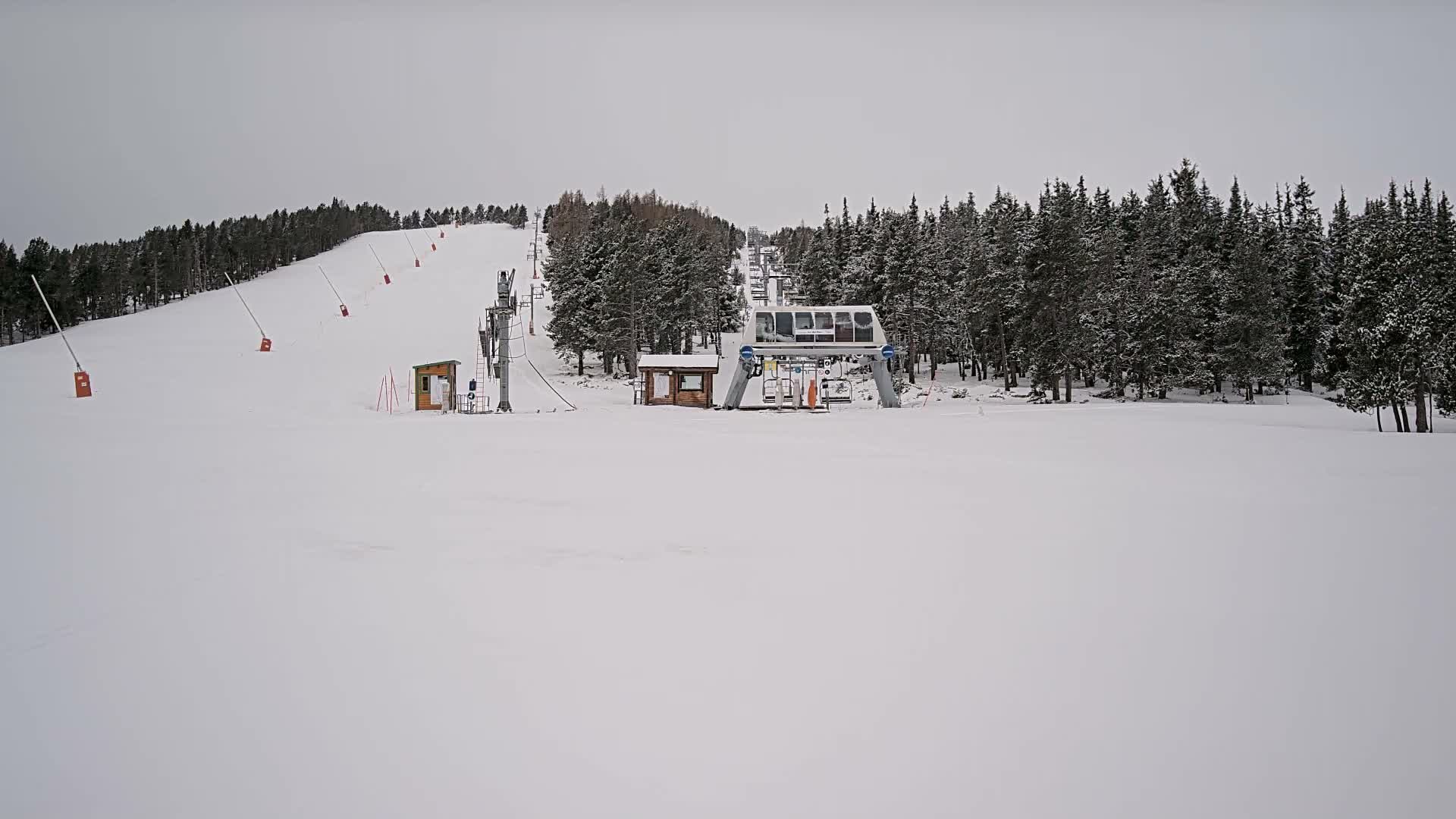 On an overcast day, a ski slope is actively being prepared with snow cannons spraying snow across a partially covered, dry grassy hill, leading up to a prominent chairlift station and ascending line amidst evergreen trees.
