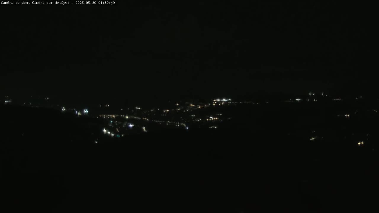 A nighttime, long-range view shows a scattering of small, faint lights from a distant town or village under a dark, cloudy sky.