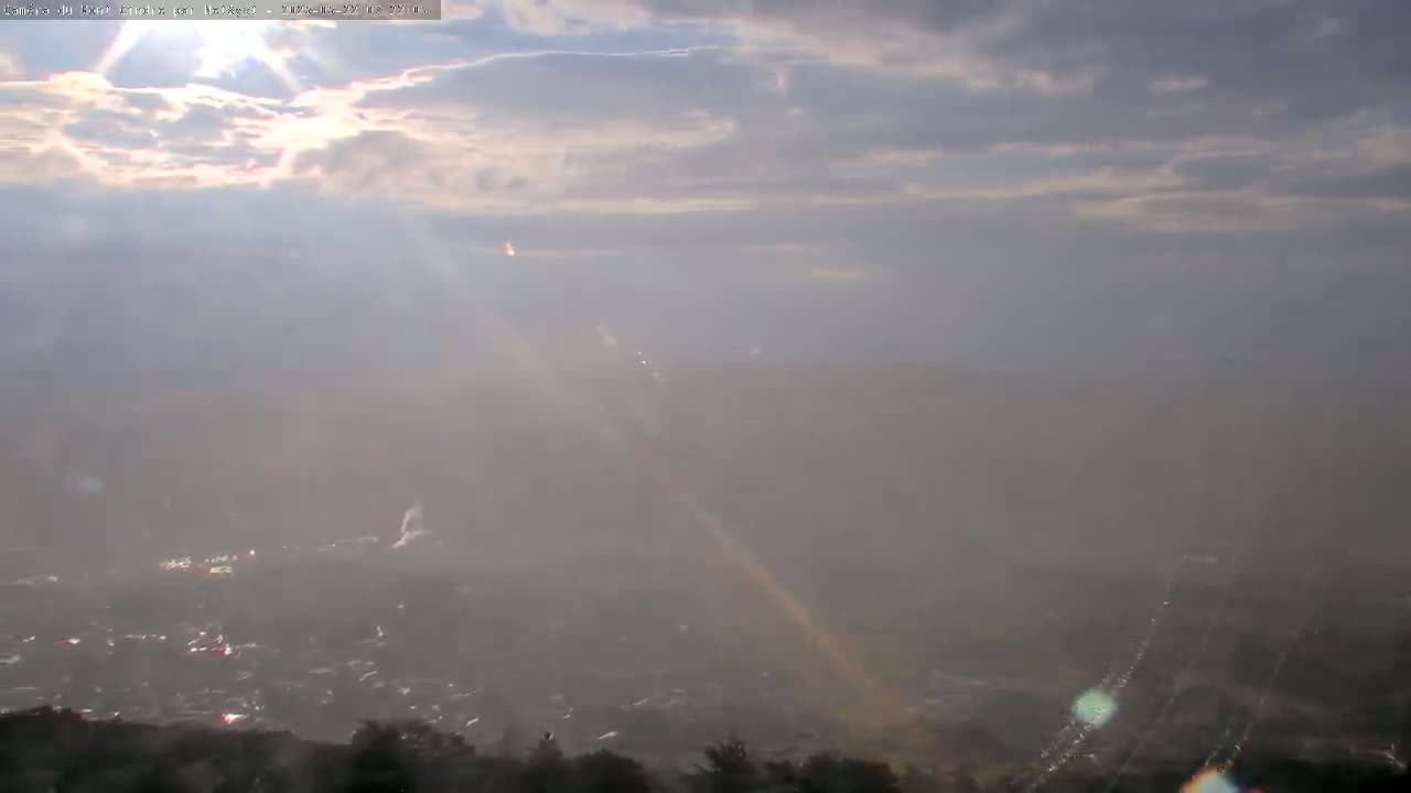 A hazy, sun-drenched view of a town below, with a partial rainbow visible amidst light rain and scattered clouds.