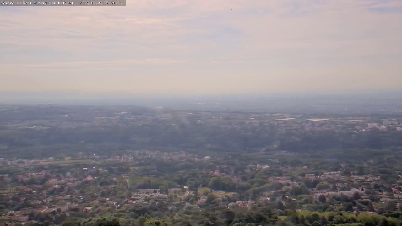 A hazy, long-distance view shows a sprawling suburban area nestled among green trees and hills extending to a distant horizon.