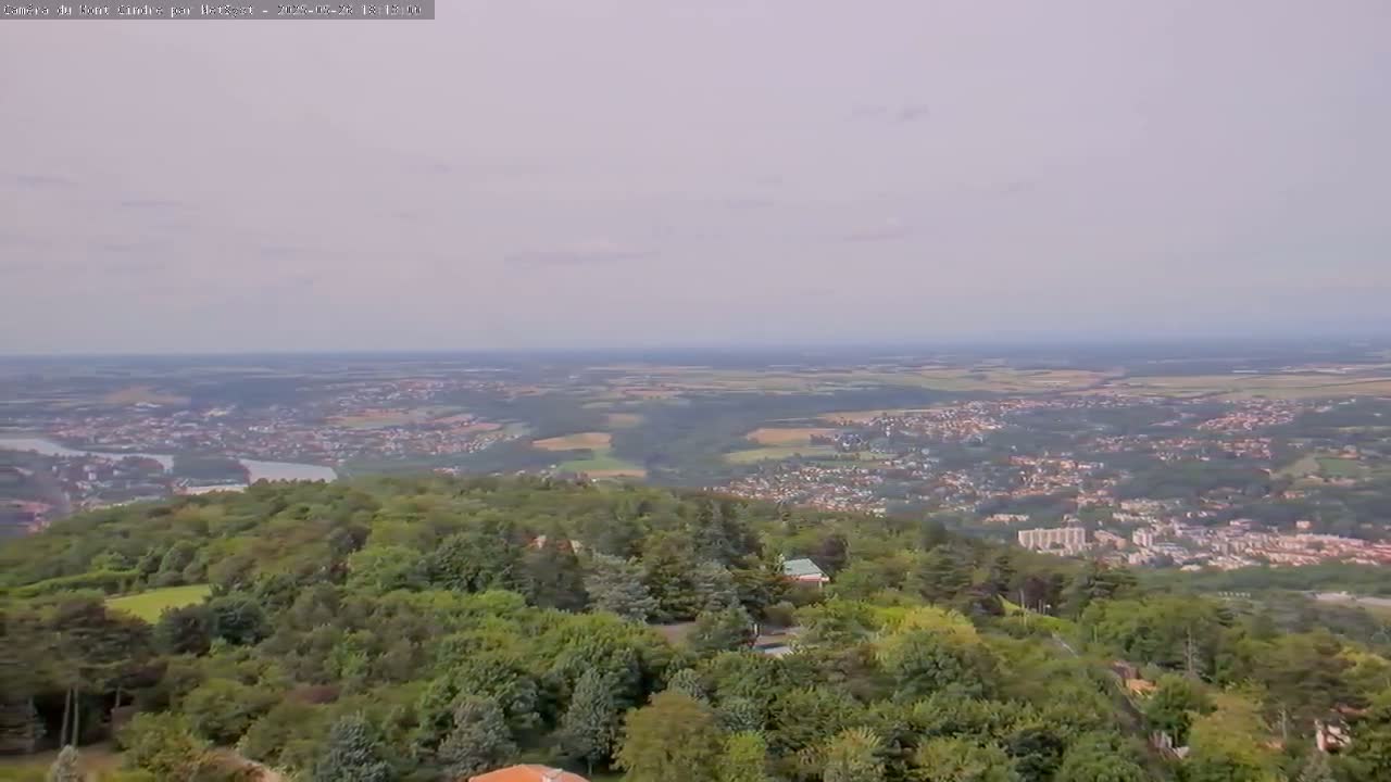 A hazy, high-angle view shows a sprawling town nestled in a valley, surrounded by lush green forests and a winding river, all under a pale sky.