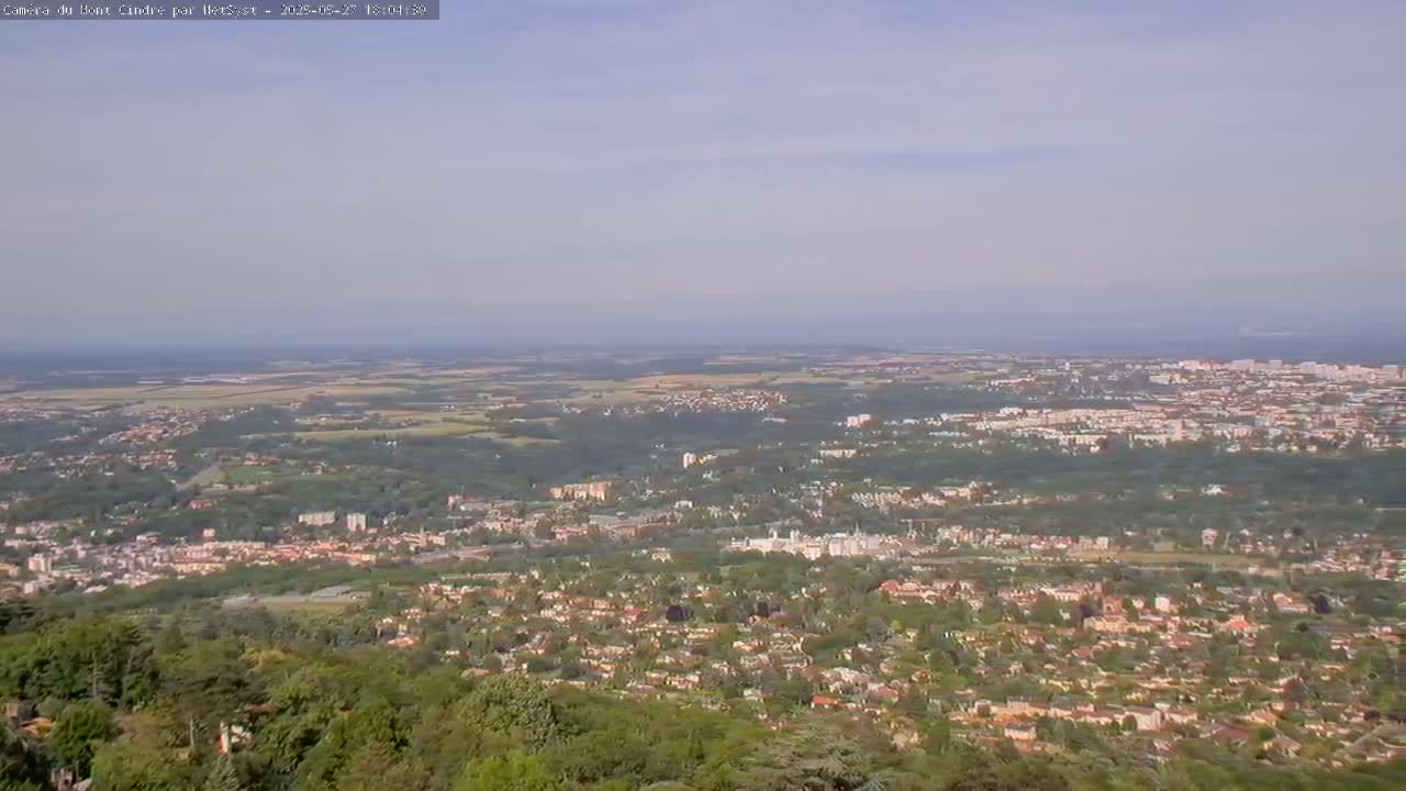 A hazy, long-range view shows a sprawling city nestled in a green landscape, extending to a distant, pale horizon under a mostly clear sky.