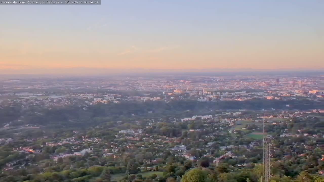 A sprawling city and its suburbs are visible from a high vantage point at sunset, under a hazy sky.