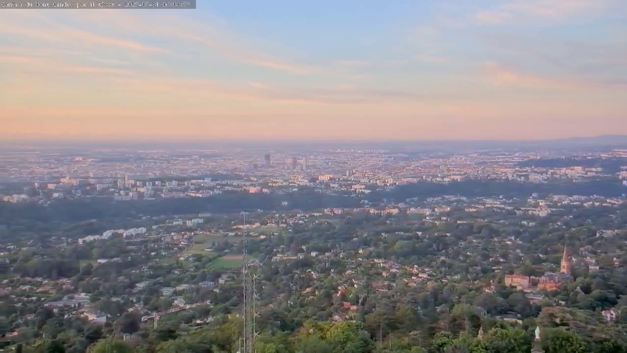 A sprawling city, bathed in the soft light of a sunset, is viewed from a high vantage point overlooking a lush, tree-covered hillside.