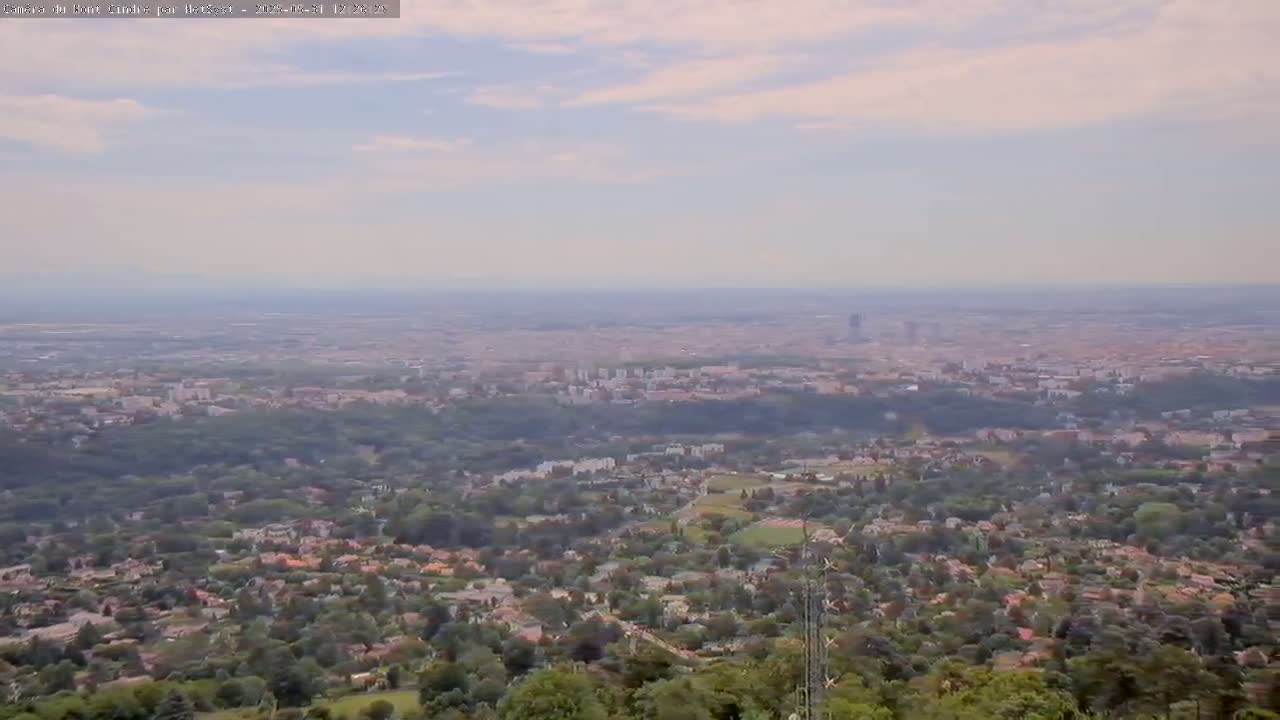 A sprawling city and its surrounding suburbs are visible from an elevated vantage point on a partly cloudy day.