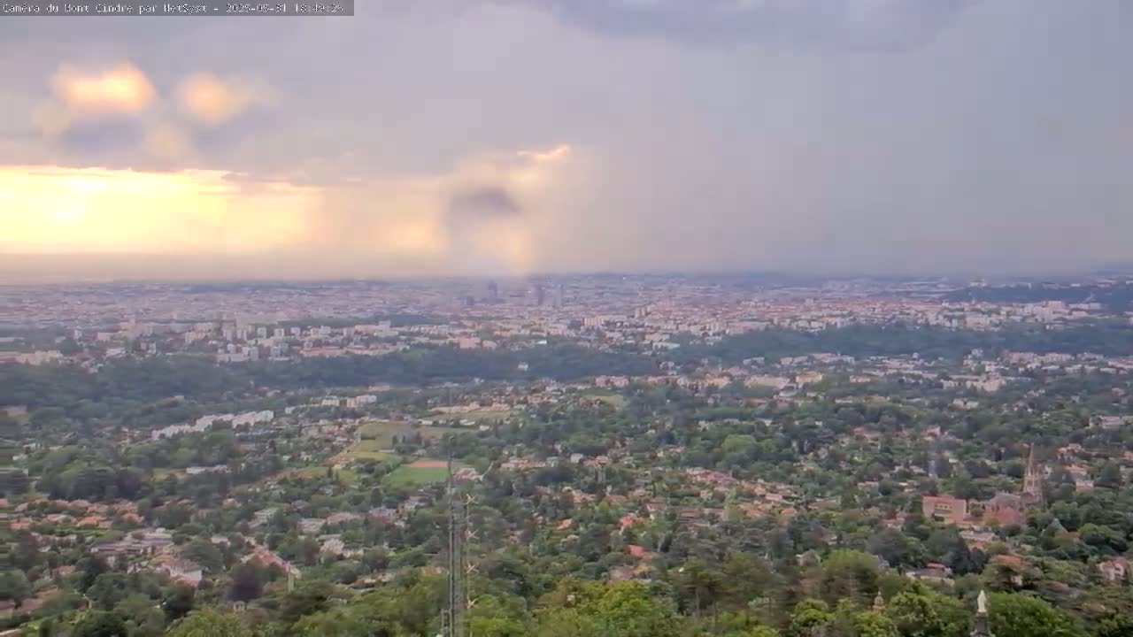 A sprawling city is visible beneath a mostly cloudy sky, with streaks of sunlight breaking through the clouds.