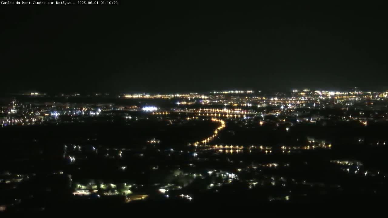 A nighttime, high-angle view shows a sprawling city illuminated by numerous lights, with a winding road visible through the darkness.