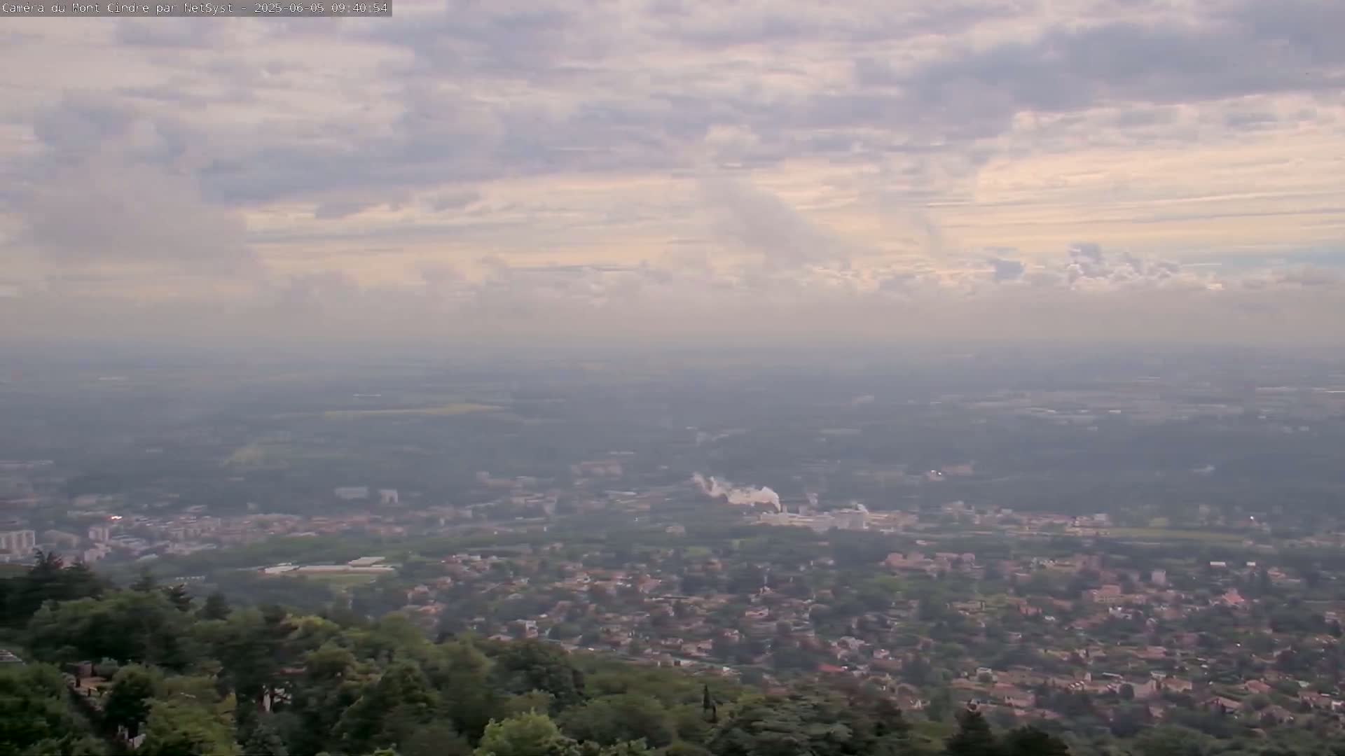 A hazy, partly cloudy sky overlooks a sprawling city nestled amongst lush green hills.