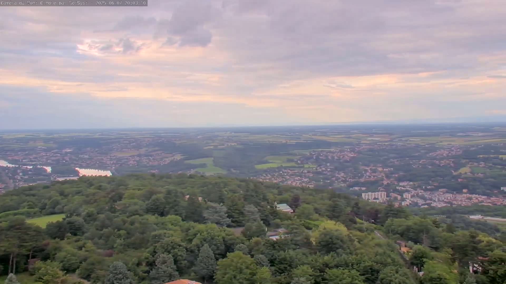 A high-angle, long shot shows a sprawling town nestled in a valley of lush green trees under a pale, cloudy sky.