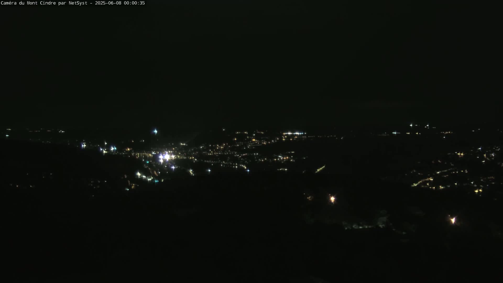 A nighttime long shot shows a city's lights spread across a dark landscape under a clear, dark sky.