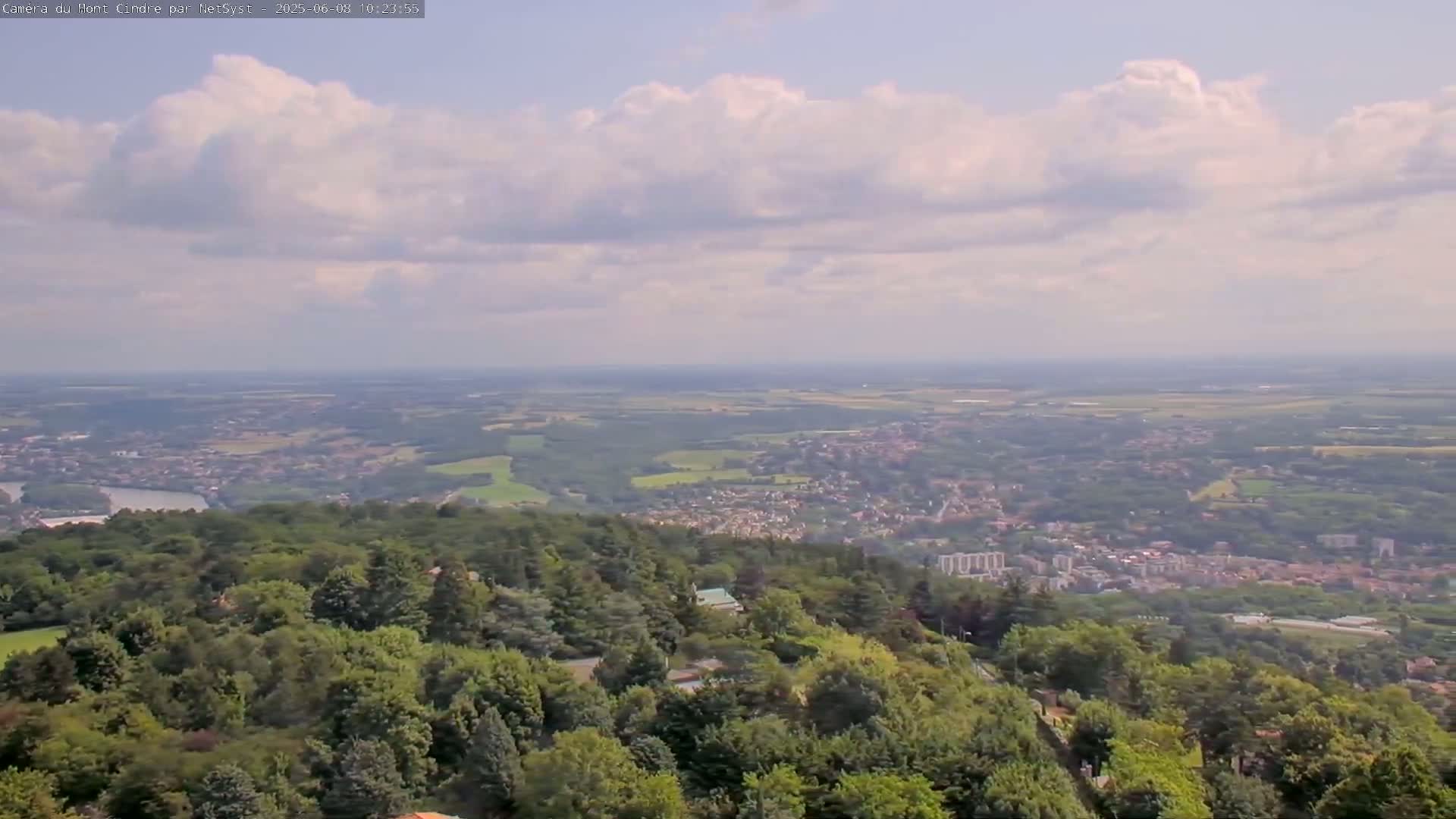 A partly cloudy sky overlooks a vast expanse of green landscape, including a town nestled amongst dense, leafy forests.