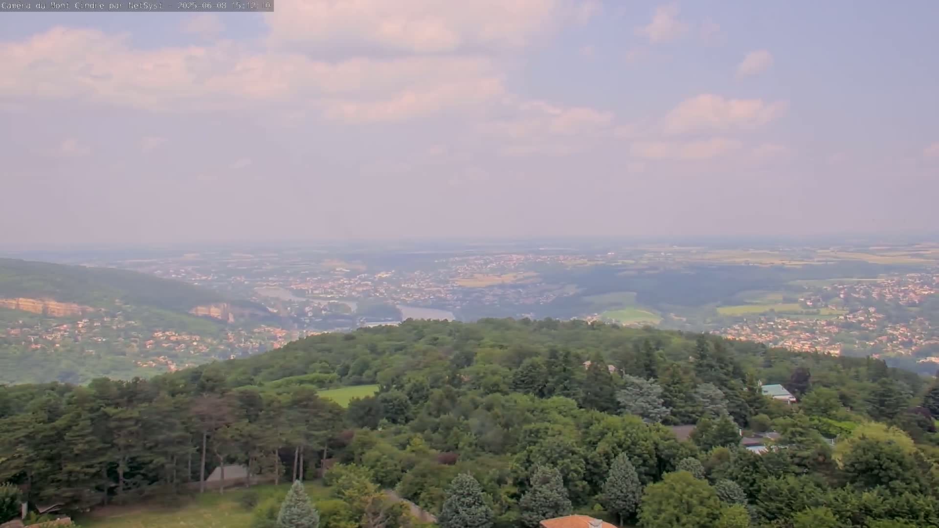 A hazy, partly cloudy day reveals a panoramic view of a vast expanse of green forest, dotted with houses and buildings, overlooking a valley with a river and a distant city.