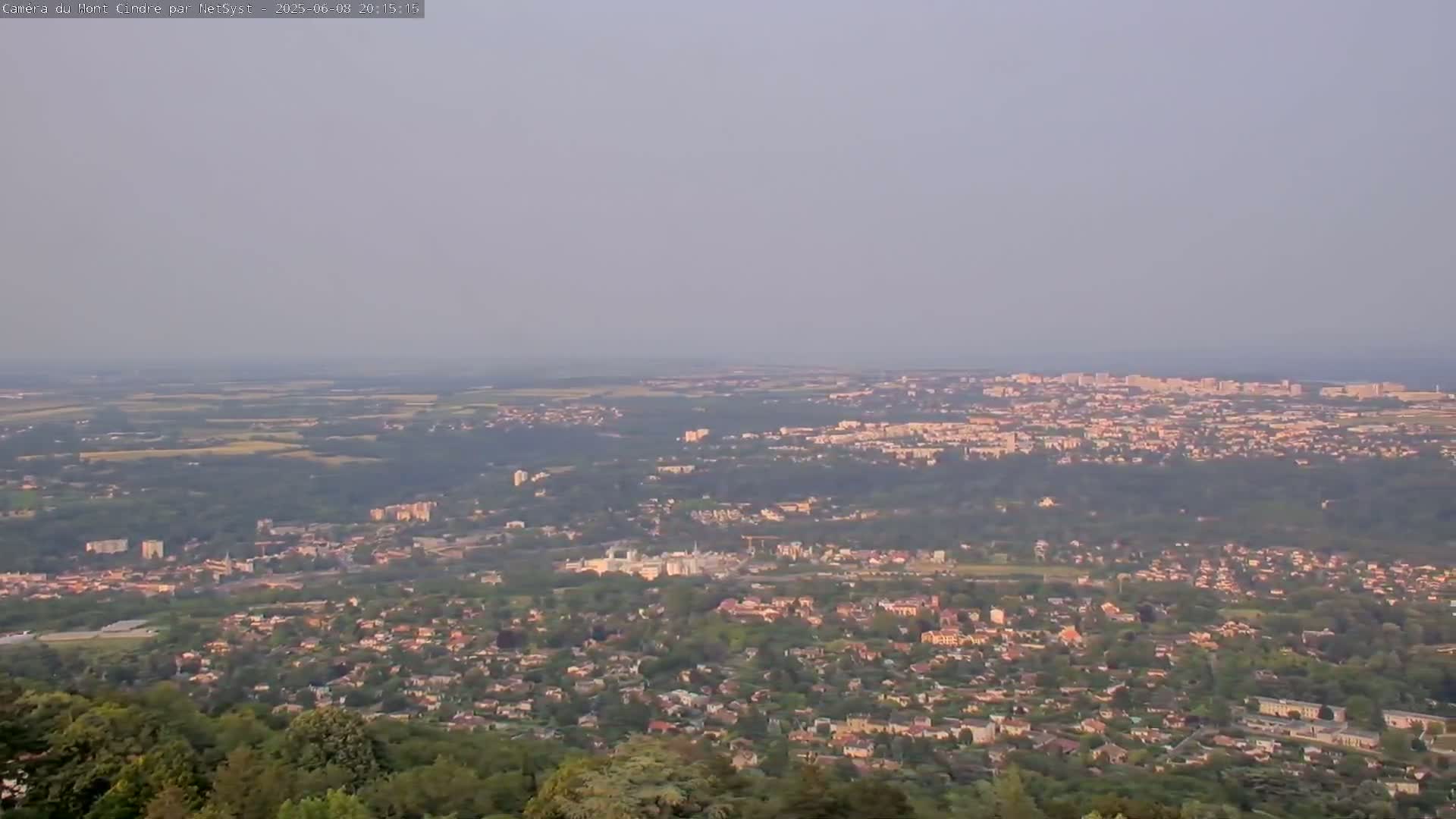 A hazy, expansive view of a city and its surrounding suburbs stretches to the horizon, under a light, muted sky.