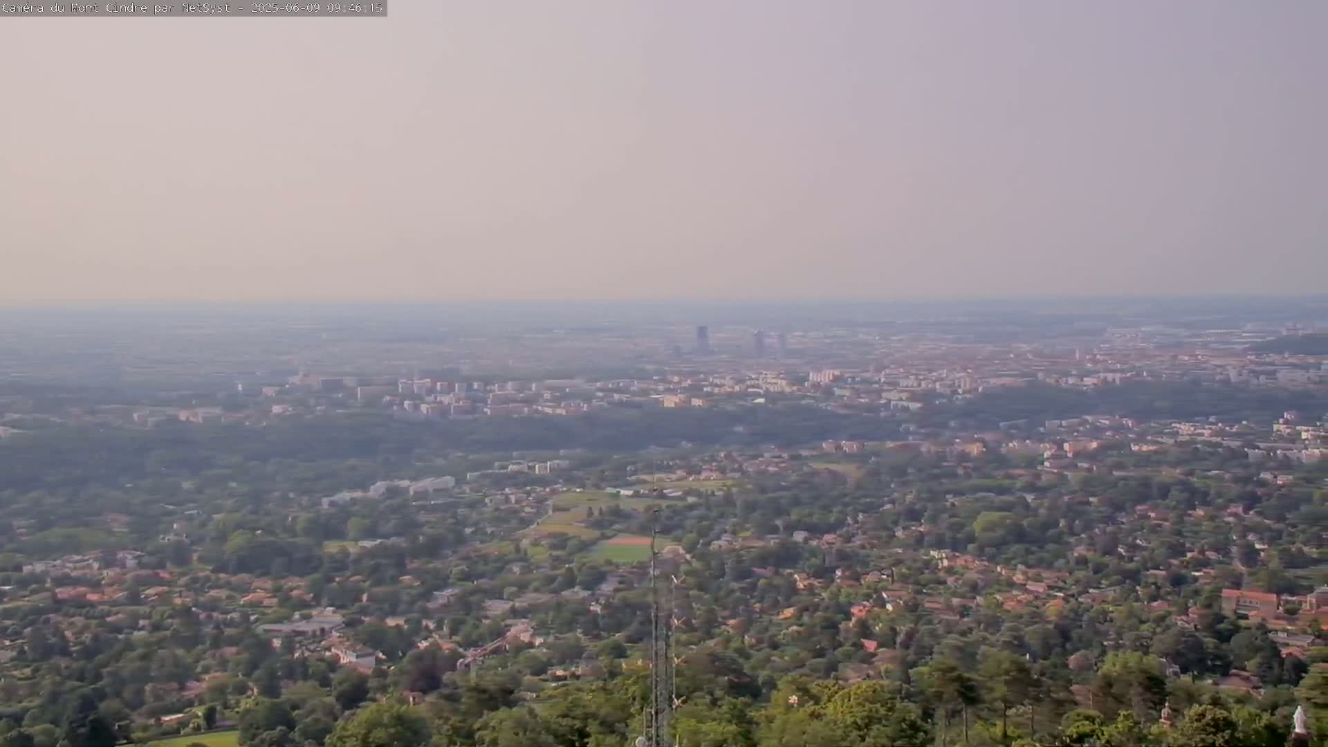 A hazy, long-range view shows a sprawling city and its suburbs nestled among green trees and hills.