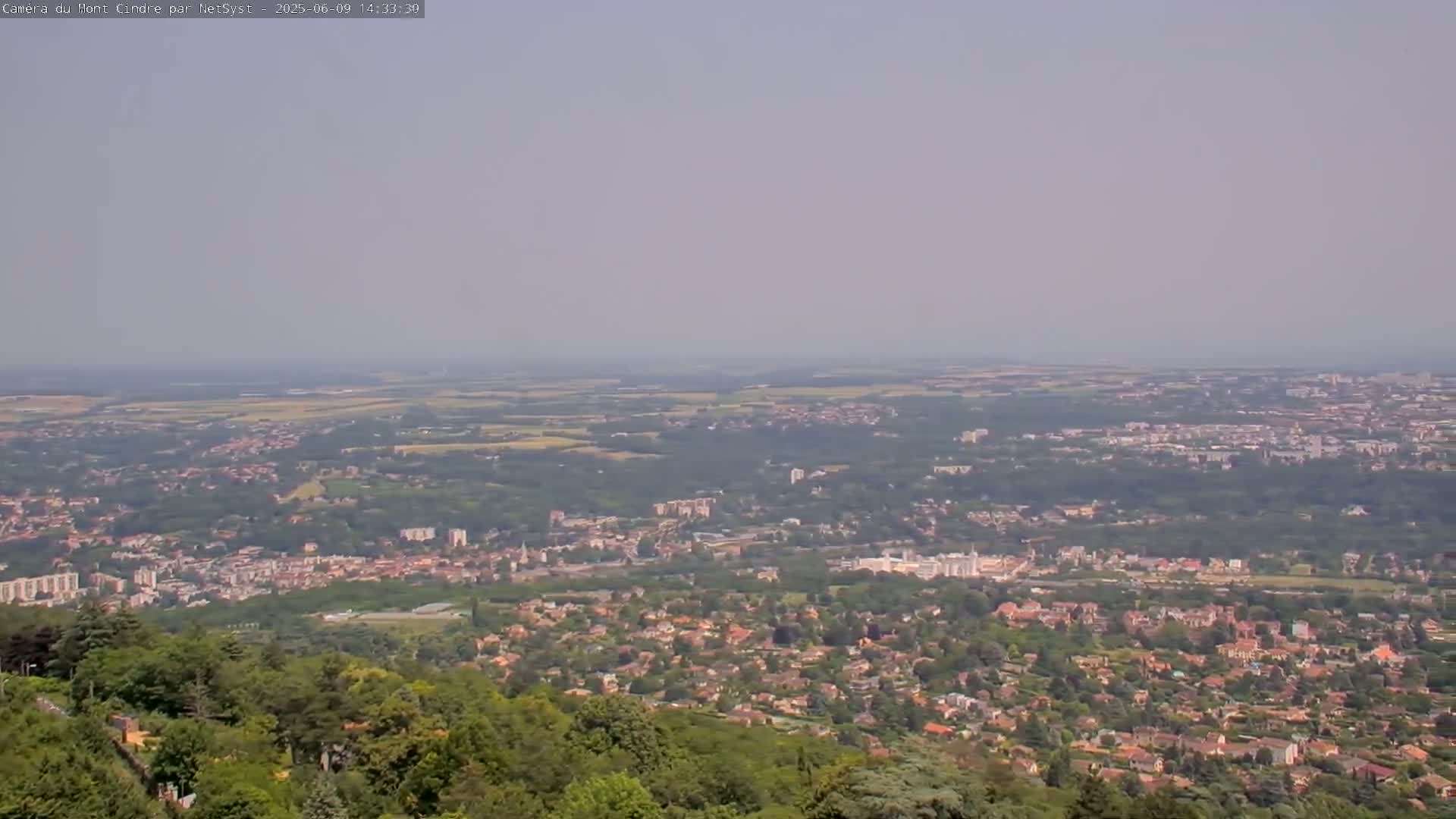 A hazy, expansive view from a high vantage point shows a sprawling town and surrounding countryside extending to the horizon.