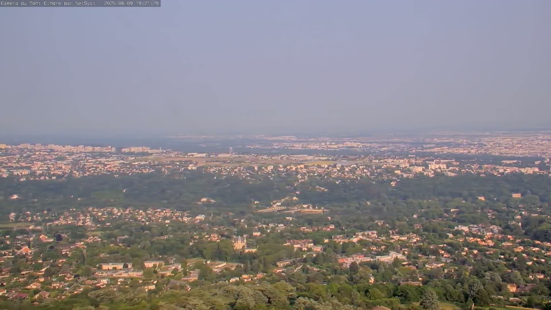 A hazy, long-range view shows a sprawling city and suburbs nestled among green trees and hills, extending to a distant coastline.