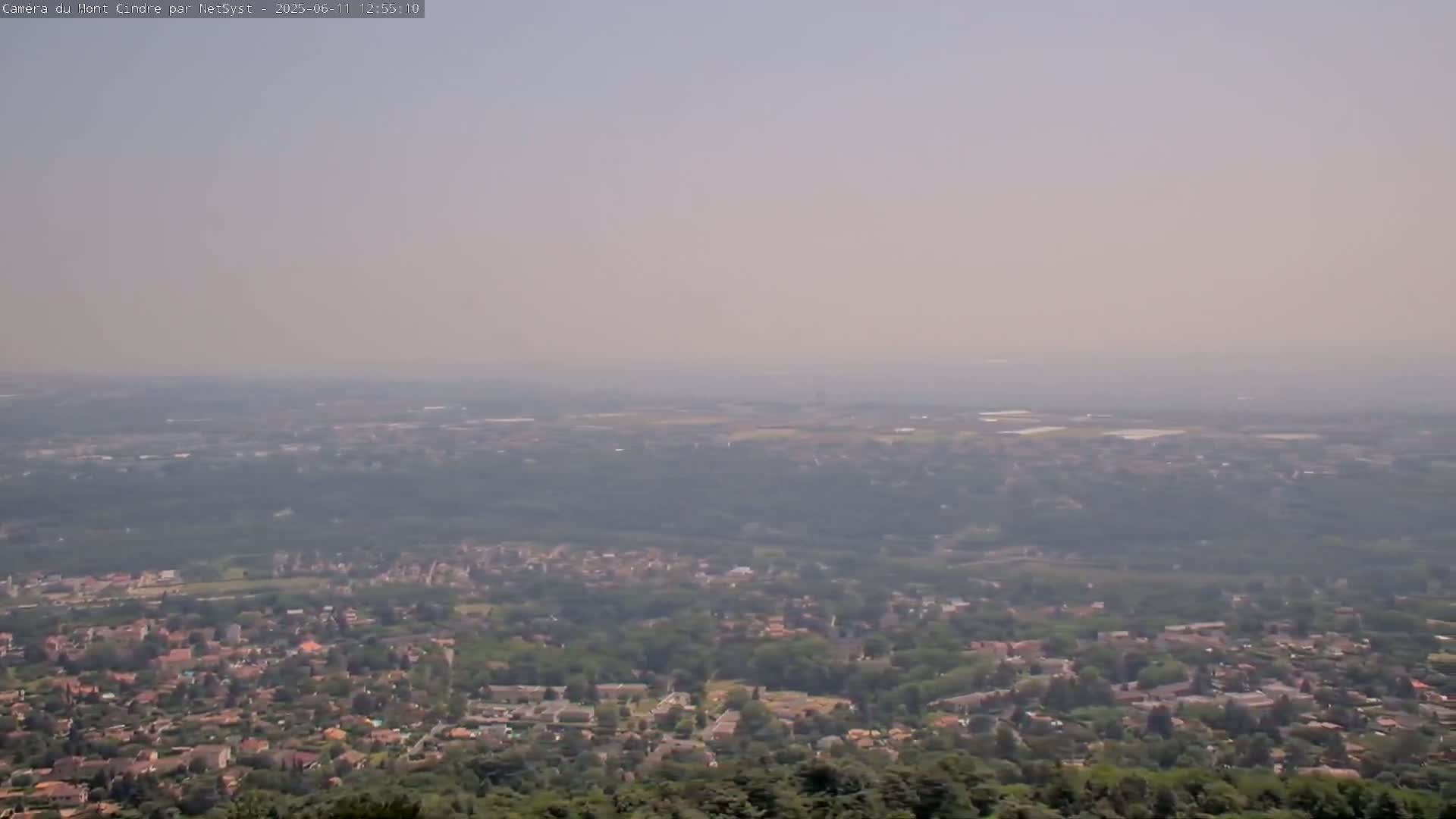 A hazy, long-distance view shows a sprawling suburban area nestled amongst green trees and fields extending to a distant horizon.