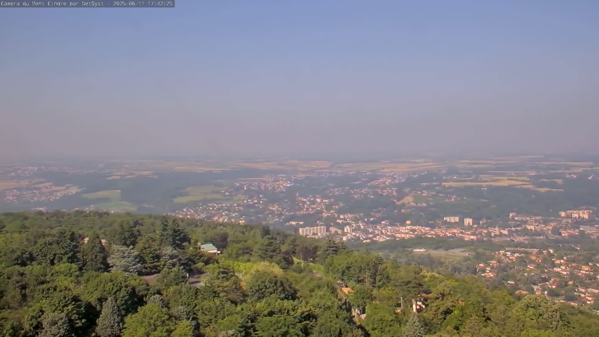 A hazy, sunny view from a tree-covered hilltop overlooks a sprawling town and surrounding fields.