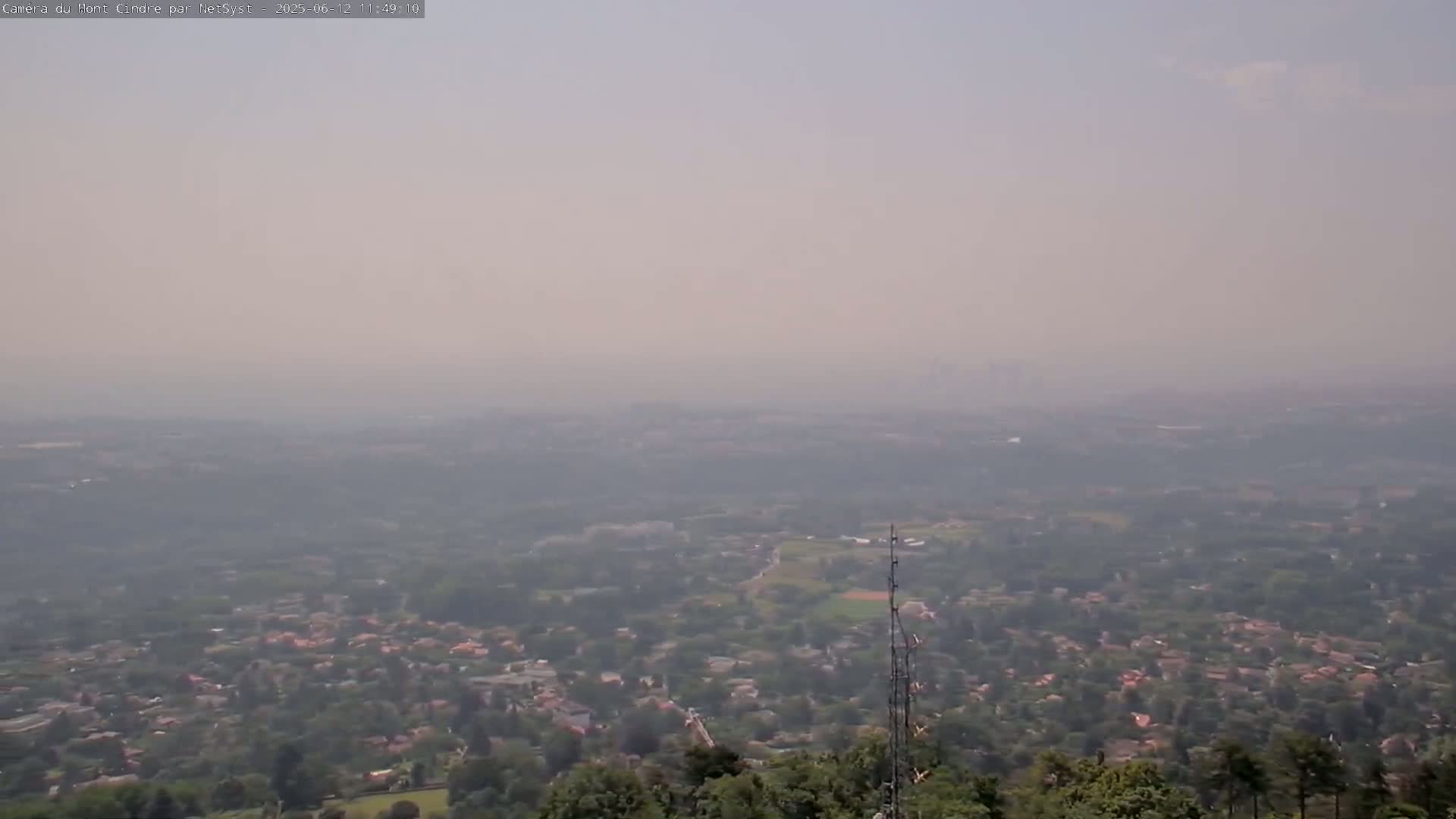 A hazy, distant view of a suburban area stretches out from a high vantage point, with a communication tower in the foreground.