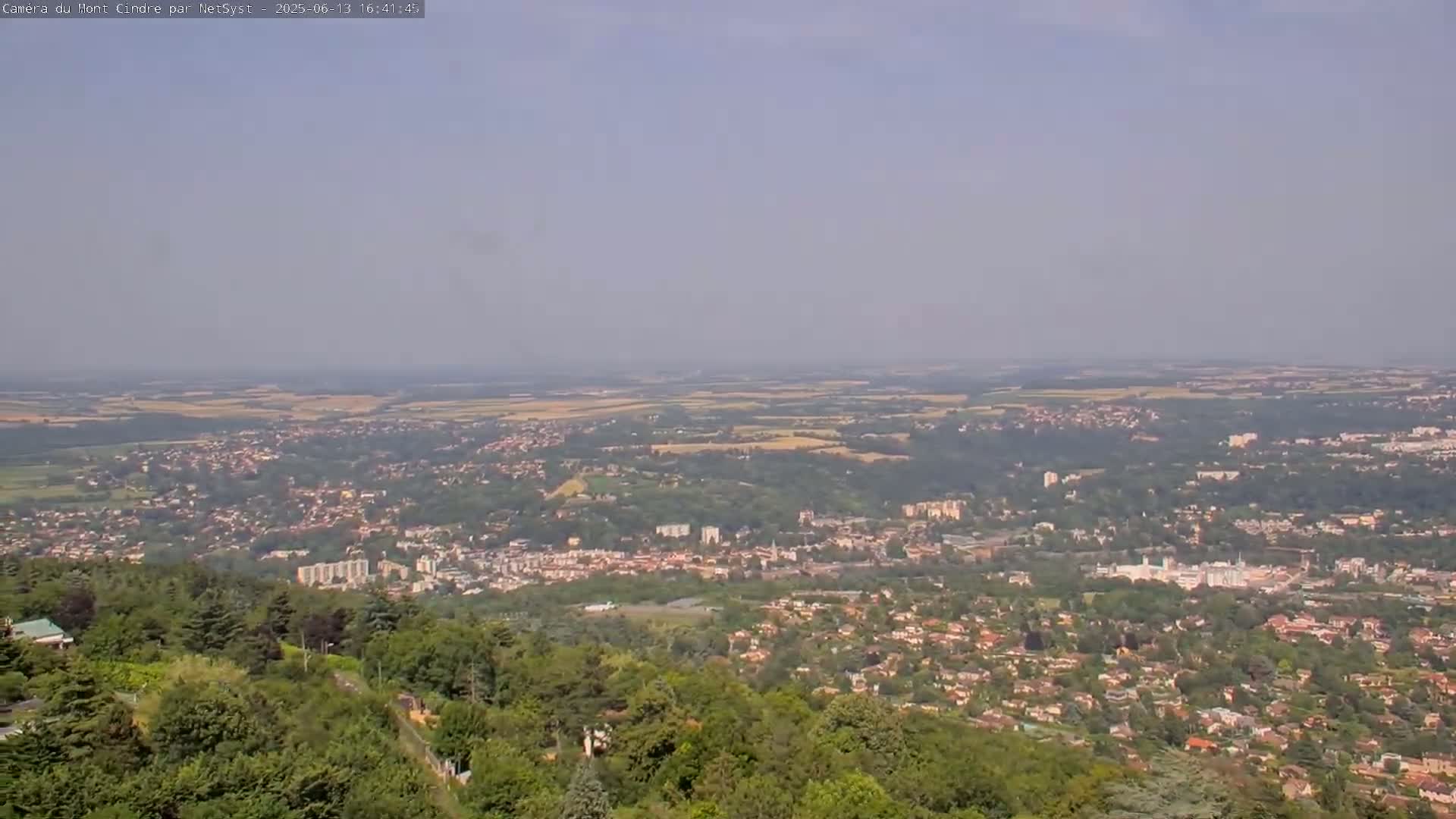 A hazy, sunny day overlooks a sprawling cityscape nestled amongst green hills and forests, extending to a vast, flat plain in the distance.