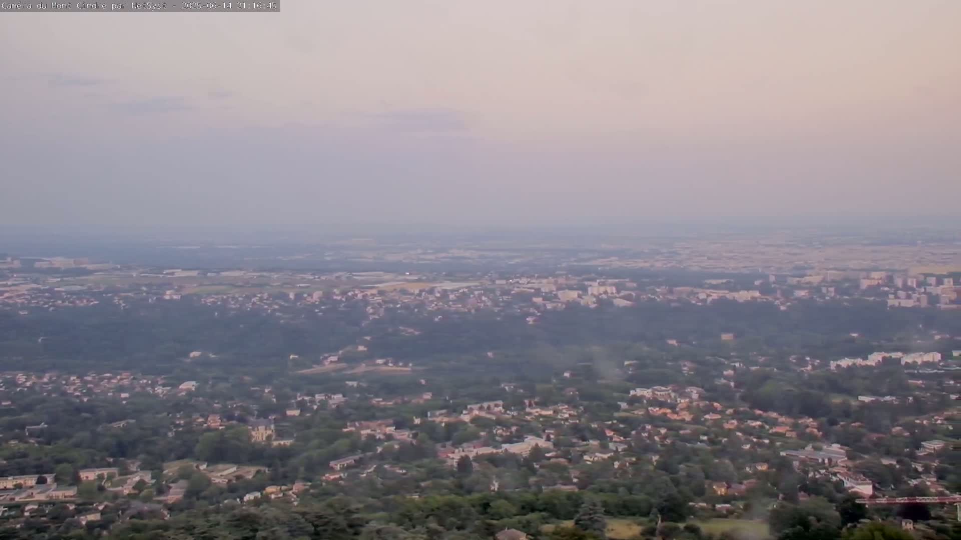 A hazy, long-distance view shows a sprawling suburban area with abundant green trees and houses, extending to a distant city under a light-grey sky.
