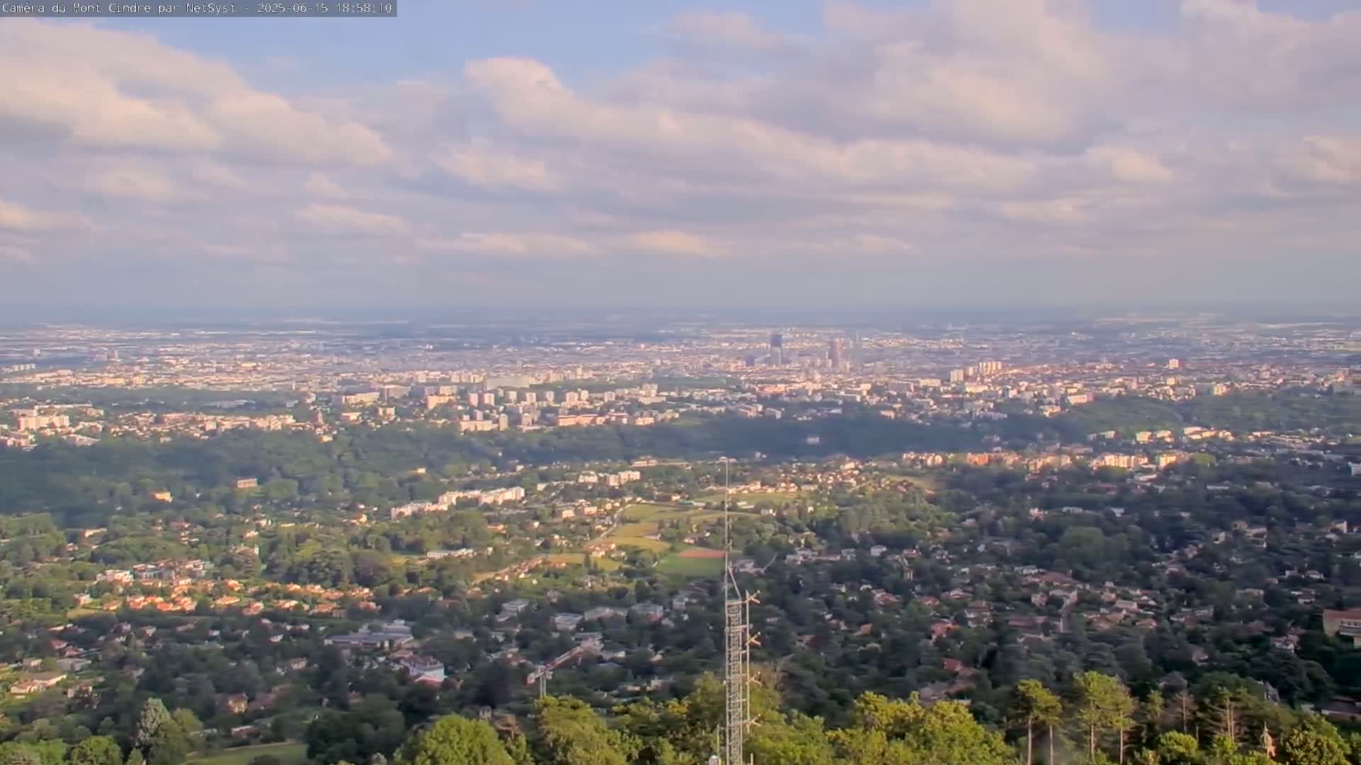 A sprawling city and its surrounding suburbs are visible from a high vantage point under a partly cloudy sky.