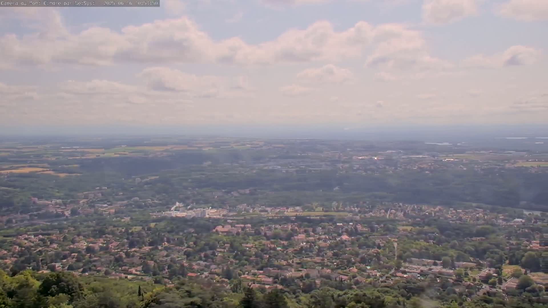 A hazy, partly cloudy day reveals a sprawling vista of a suburban area nestled among green hills and forests, extending to a distant horizon.
