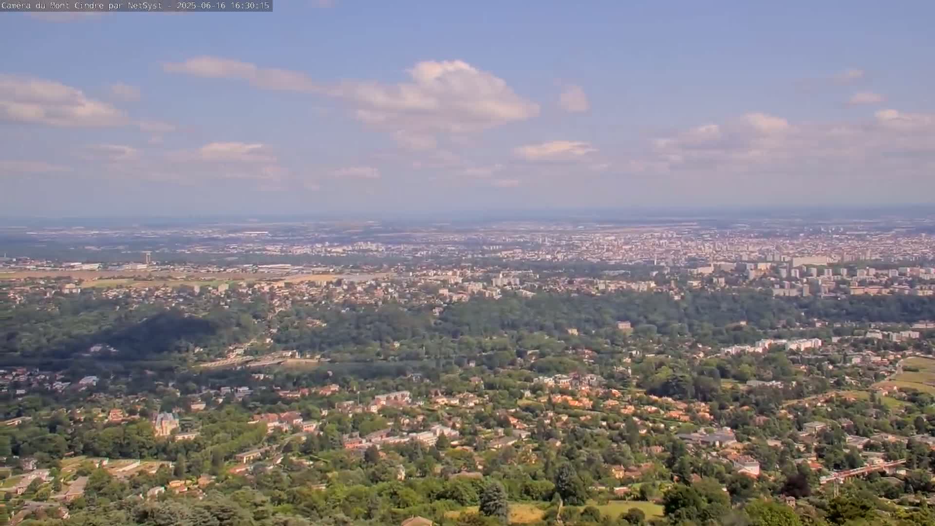A sprawling city and its surrounding suburbs are visible from a high vantage point under a partly cloudy sky.