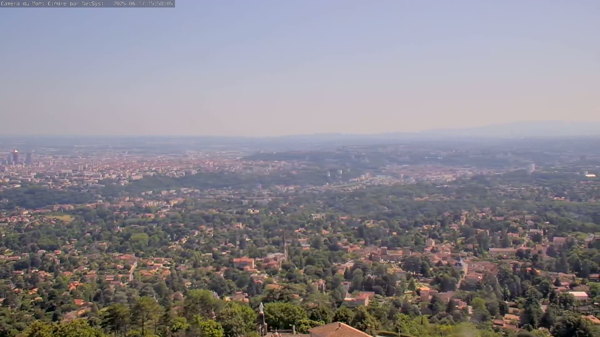 A hazy, long-distance view shows a sprawling city nestled amongst green hills and trees, extending to distant mountains under a clear, light-blue sky.
