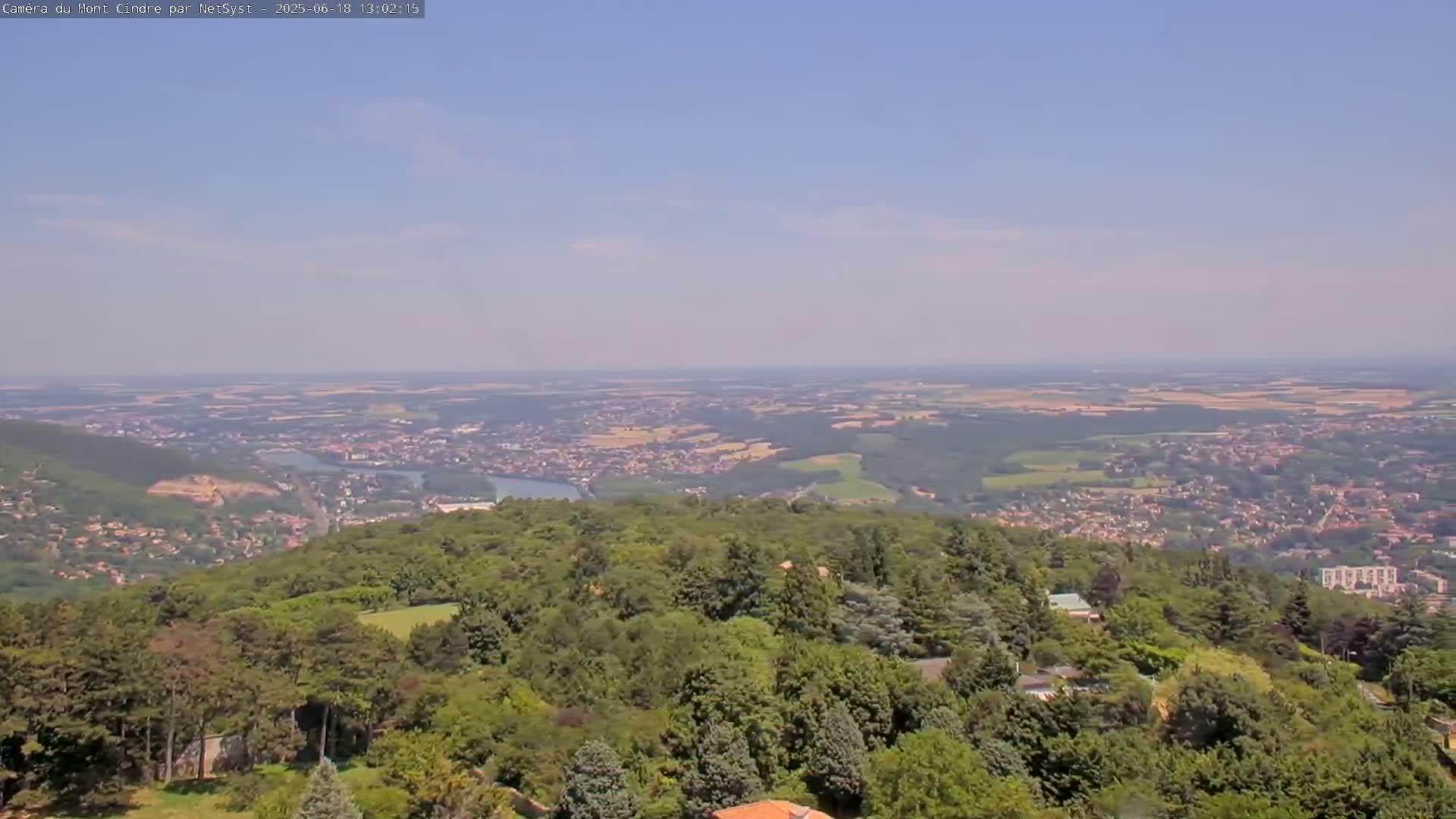 A panoramic view shows a vast expanse of green landscape, including a town nestled in a valley below, under a clear, light blue sky.