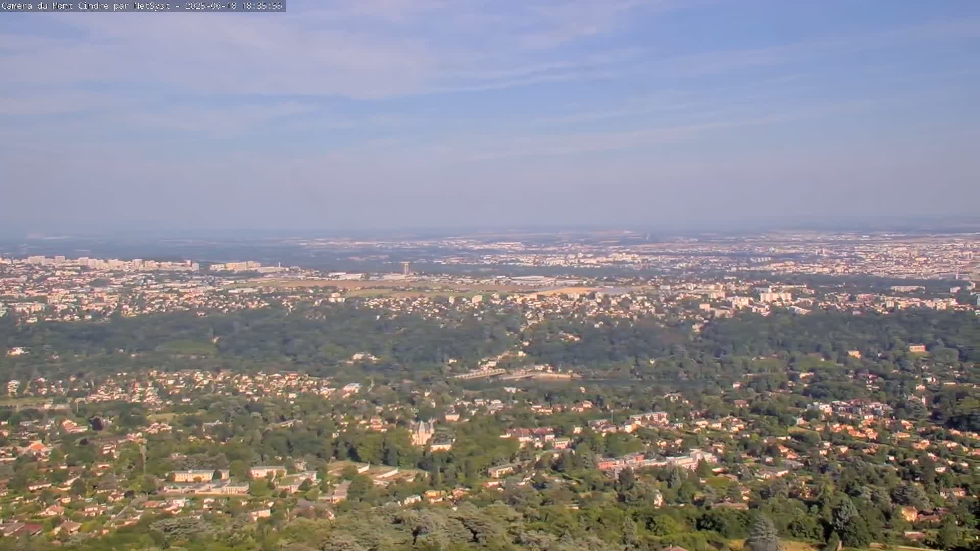 A sprawling suburban landscape with a mix of houses and trees stretches out under a mostly clear, hazy sky.