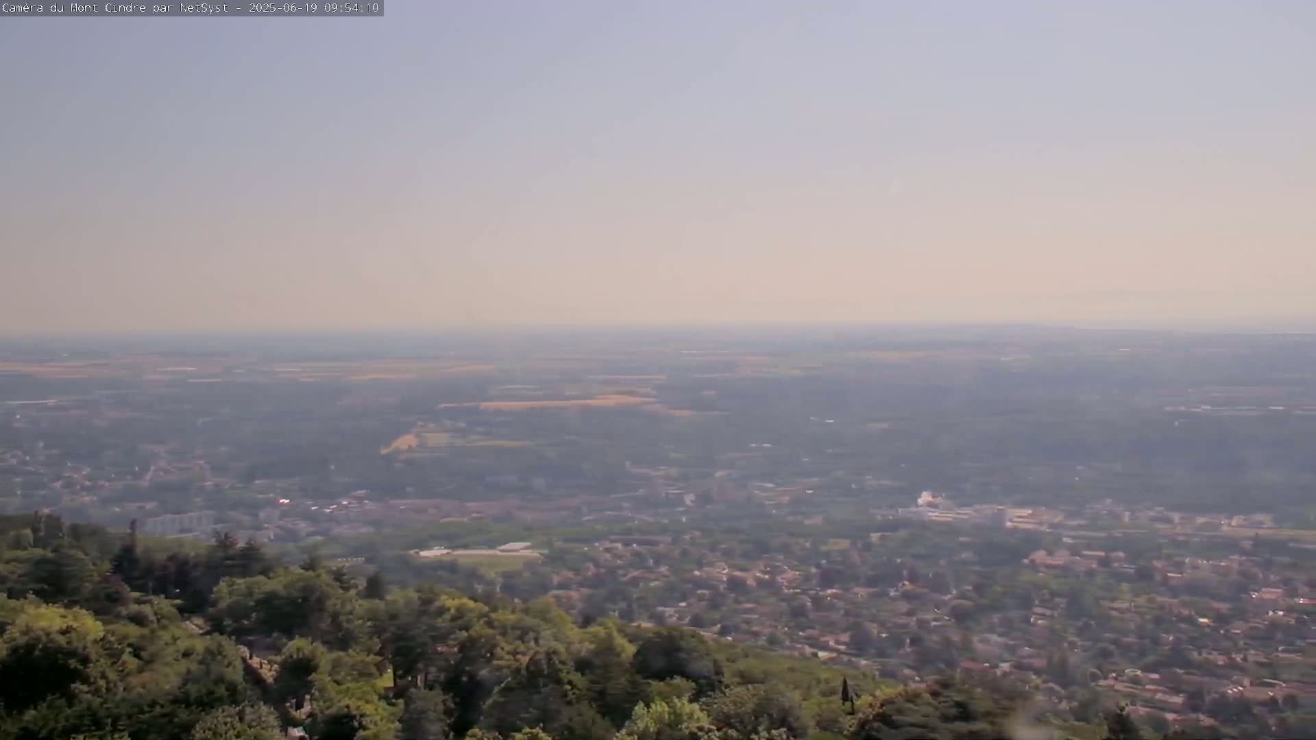 A hazy, long-range view shows a sprawling town and surrounding countryside from a high vantage point, with green trees in the foreground.
