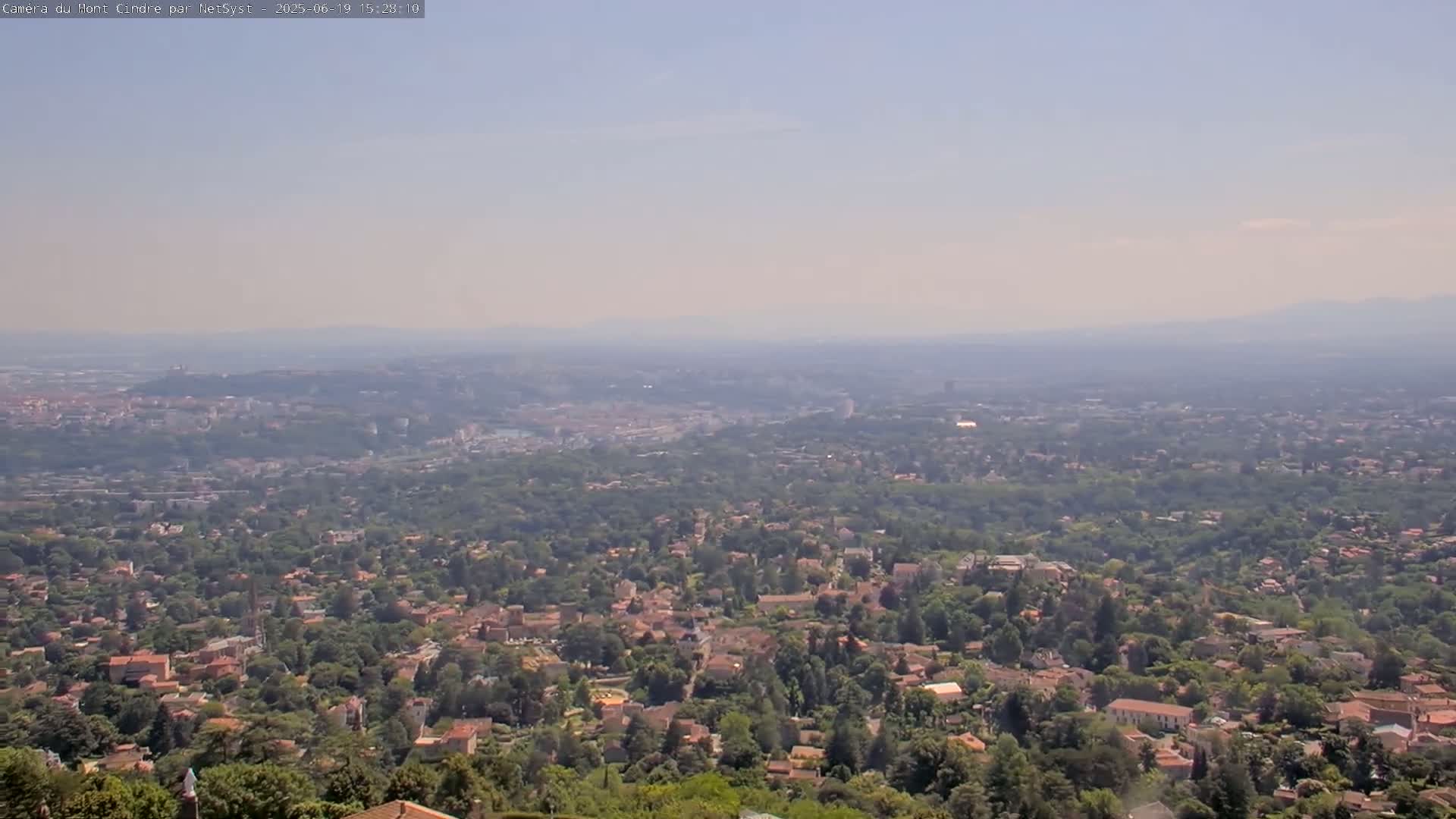 A hazy, sunny day reveals a sprawling suburban landscape dotted with houses and trees, stretching to a distant city on the horizon.