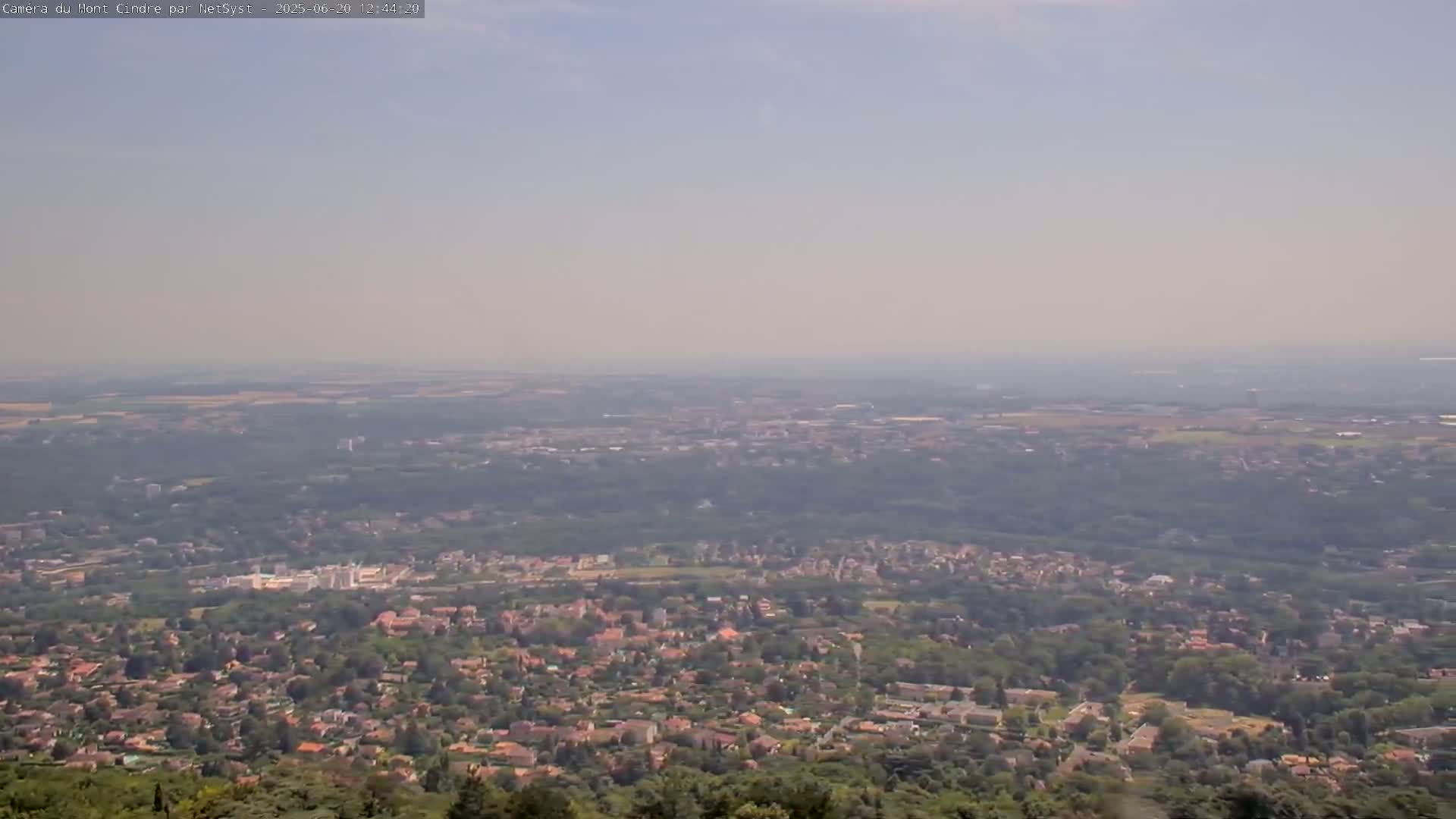 A hazy, long-distance view shows a sprawling residential area nestled among green trees and fields, extending to a distant horizon under a pale sky.