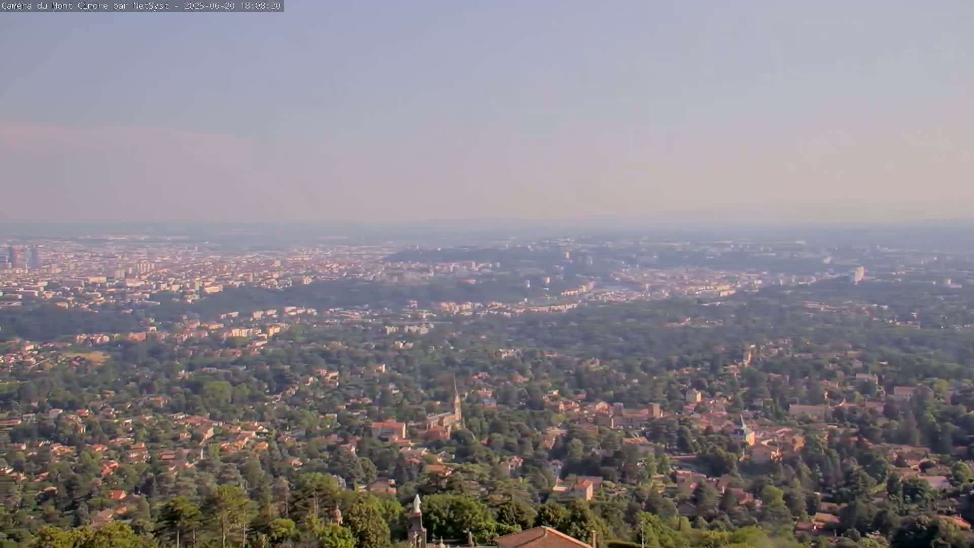 A hazy, long-distance view shows a sprawling city surrounded by lush green hills and forests under a clear, light-blue sky.