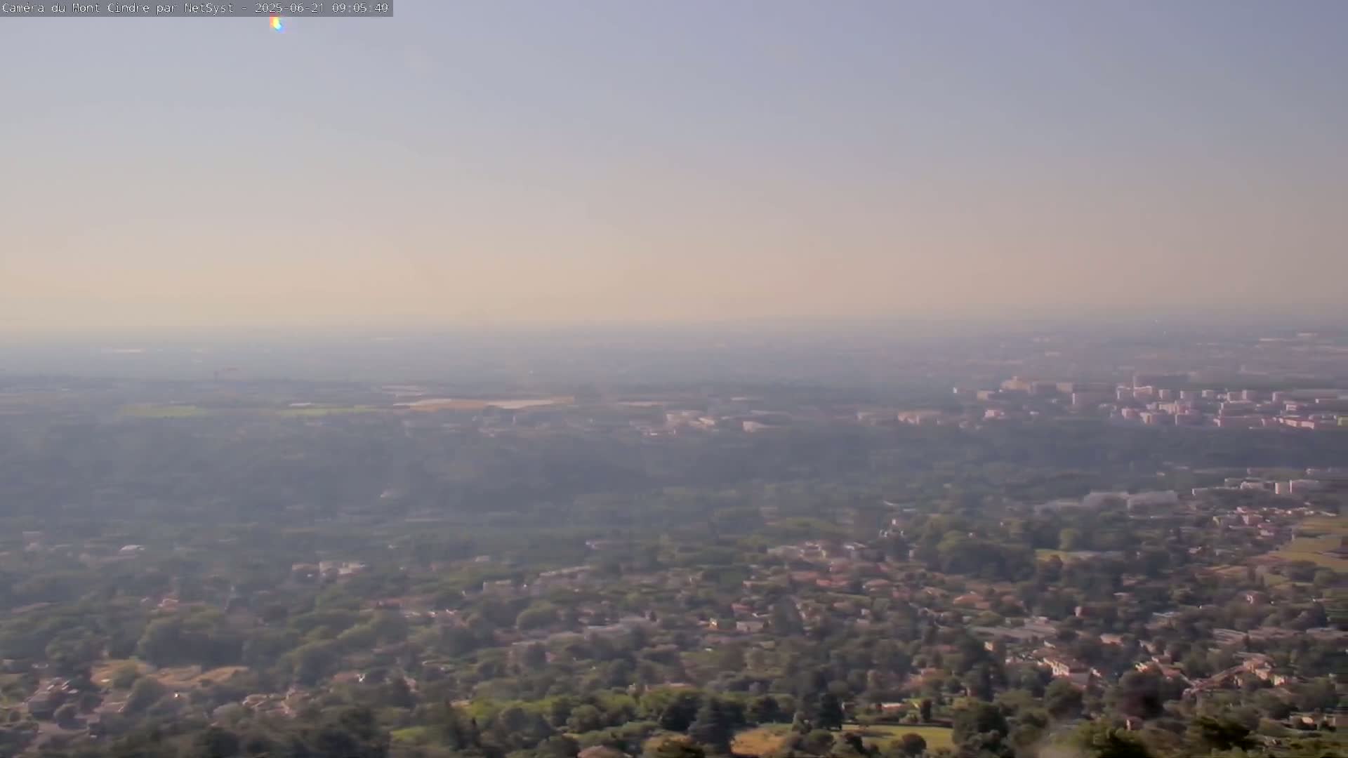 A hazy, long-range view shows a sprawling suburban area with patches of trees and a city on the horizon under a light-colored sky.