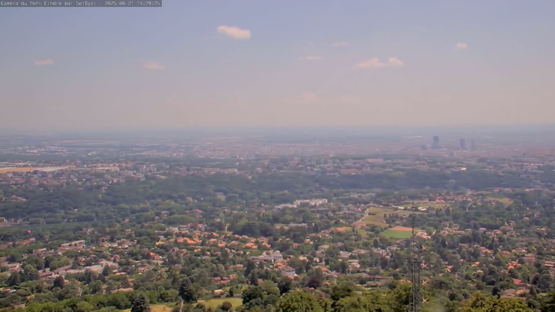 A hazy, sunny day reveals a sprawling suburban landscape nestled amongst green trees, with a distant city skyline visible on the horizon.