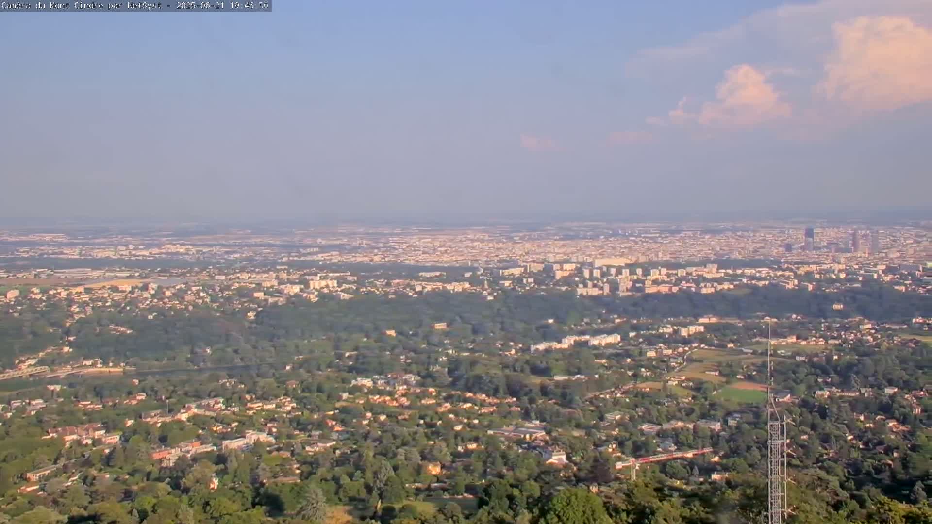 A hazy, partly cloudy day reveals a sprawling cityscape nestled amongst lush green forests and residential areas, viewed from a high vantage point.