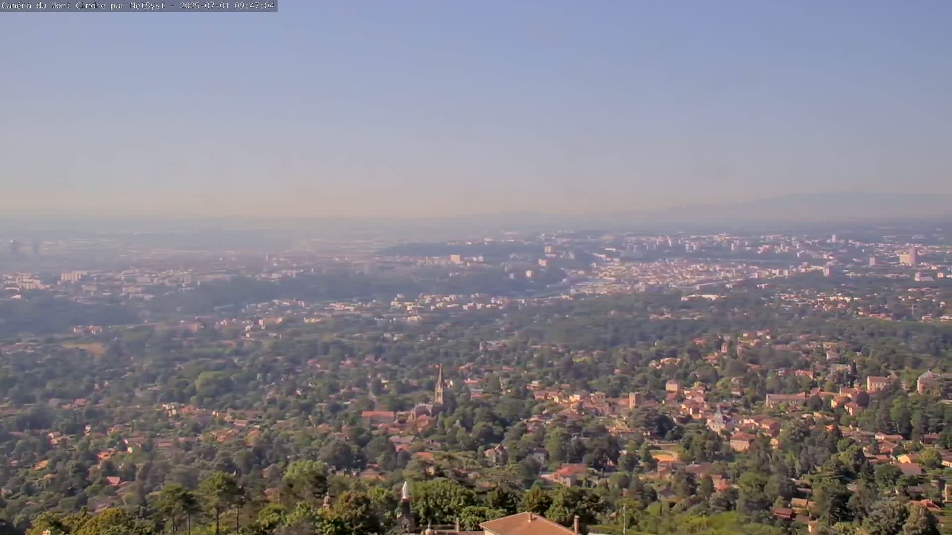 A hazy, distant city sprawls across a vast landscape of lush green trees and residential areas.