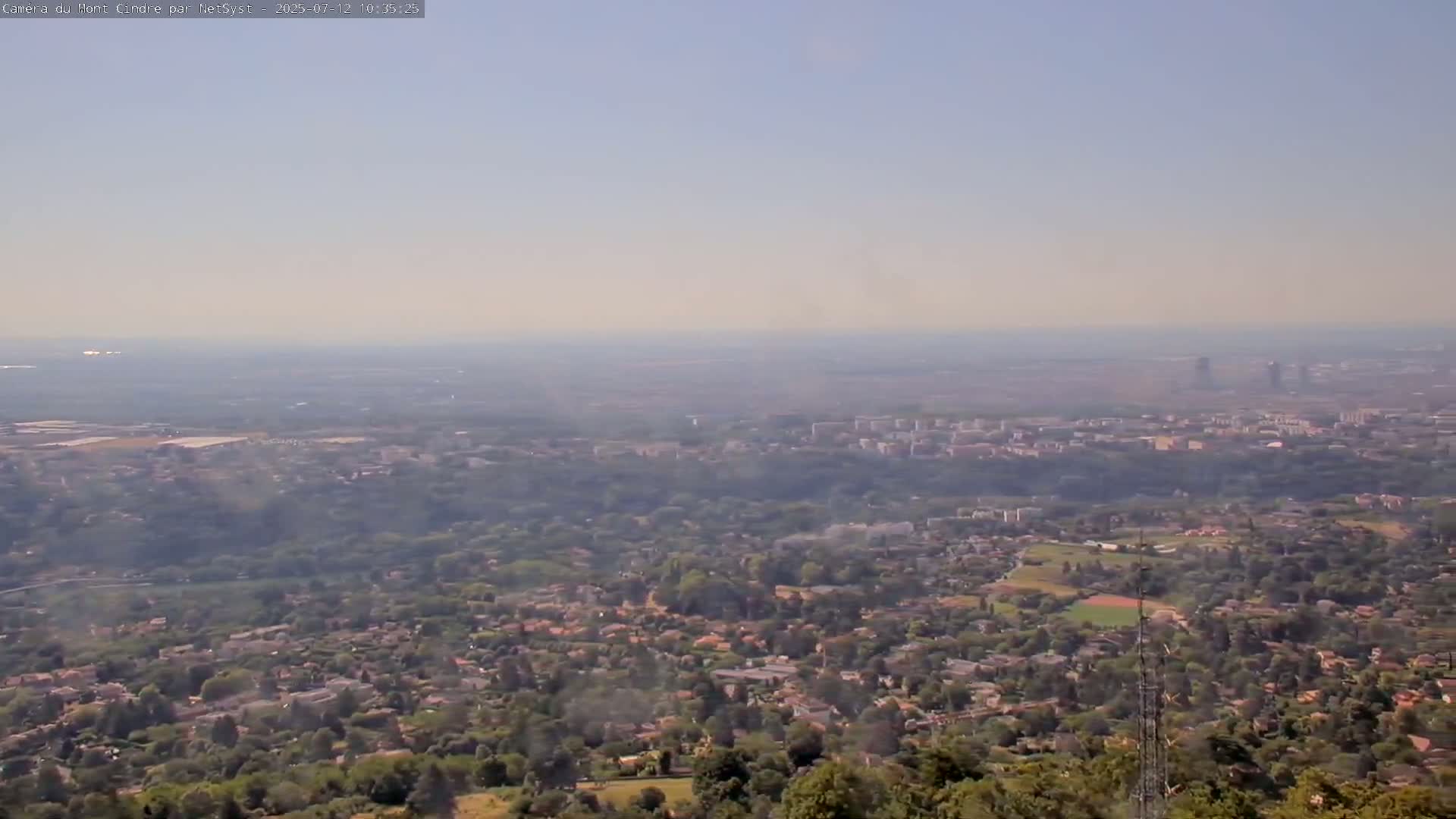A hazy, long-range view shows a sprawling suburban area nestled amongst green trees, extending to a distant city skyline under a light, clear sky.